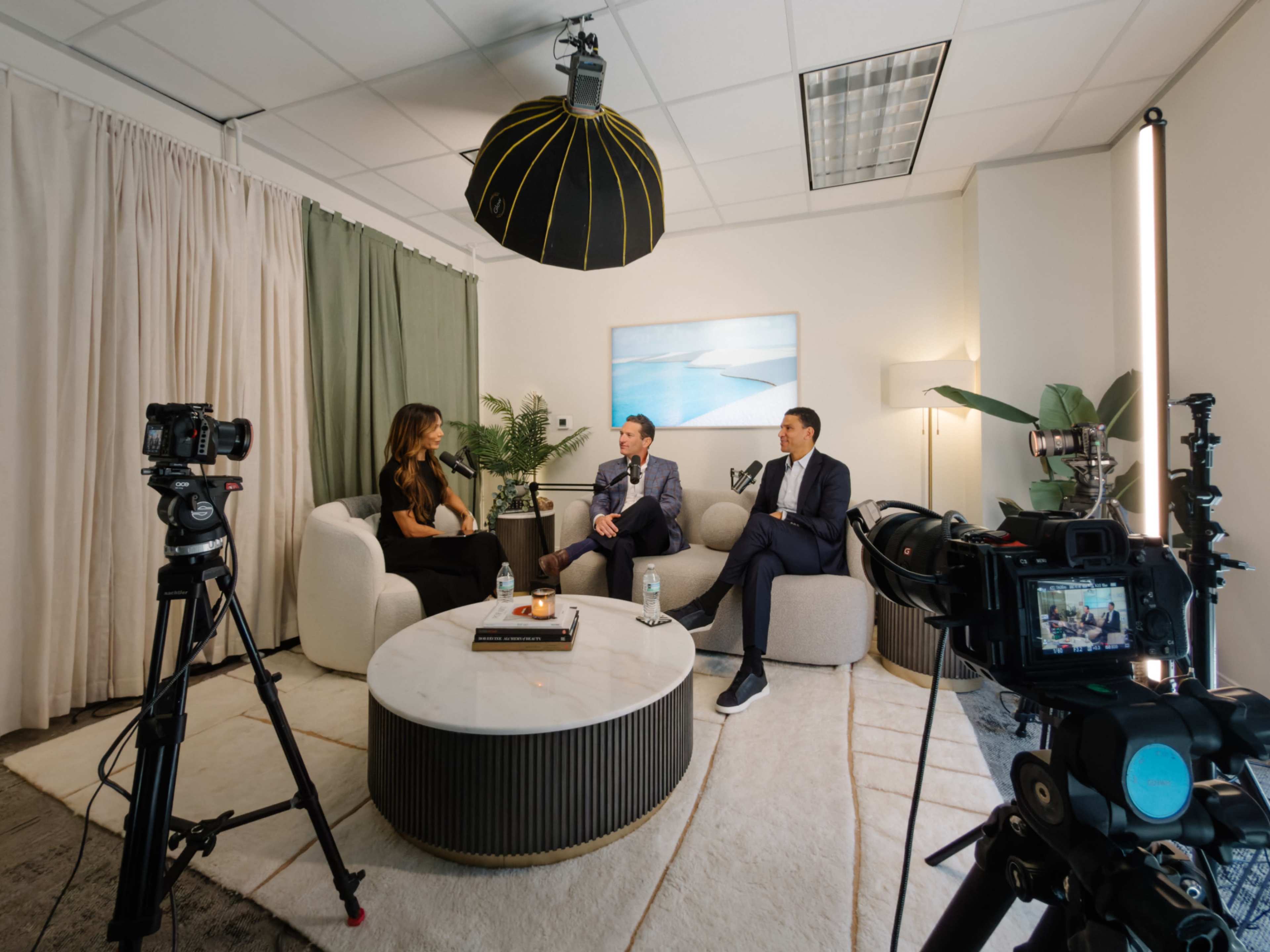 Two men in suits and a woman are seated on a sofa in a well-lit interview setting, surrounded by cameras and a large round table.