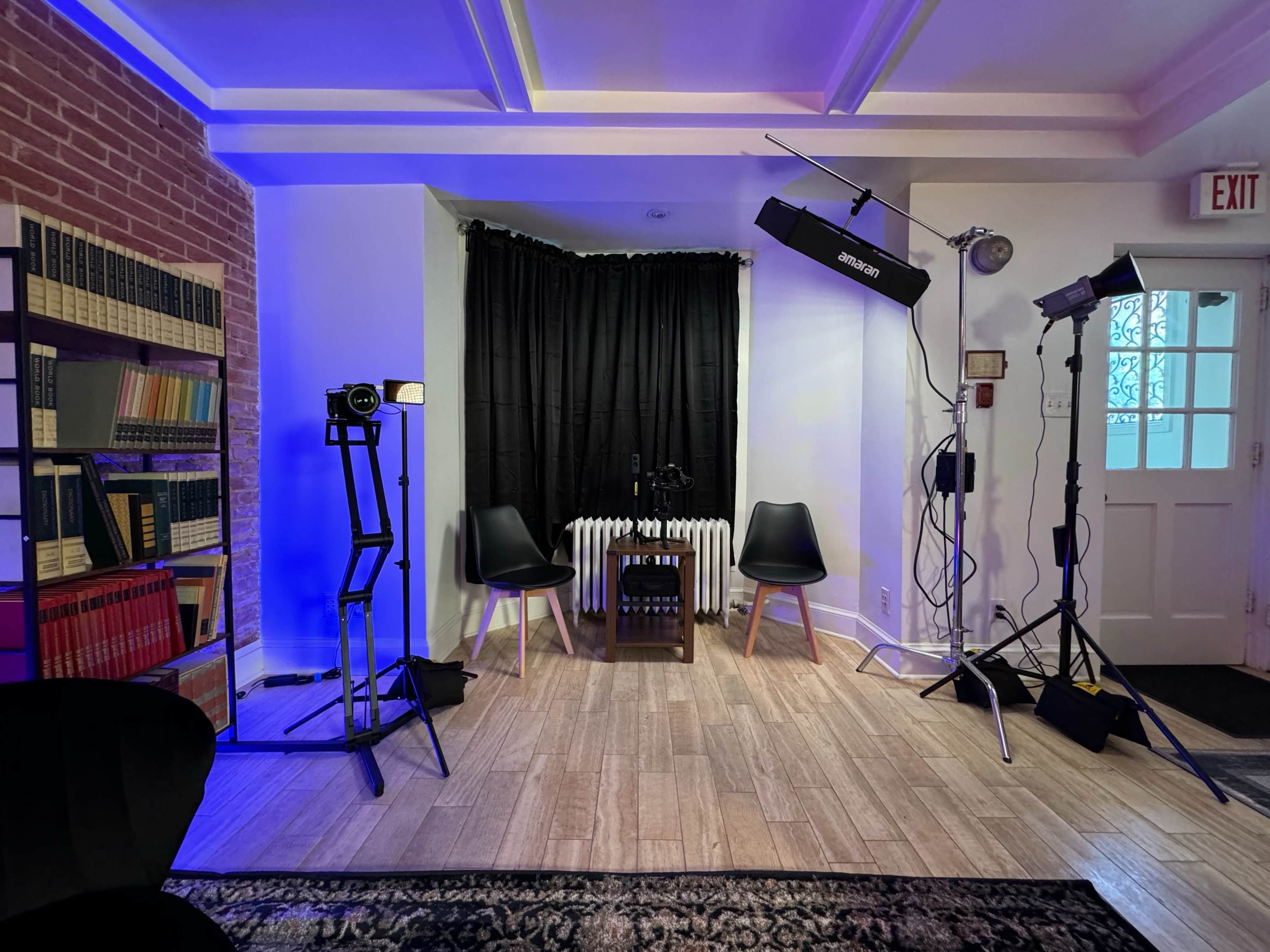A small studio setup featuring two chairs, a table, and several lighting equipment arranged around a black curtain.