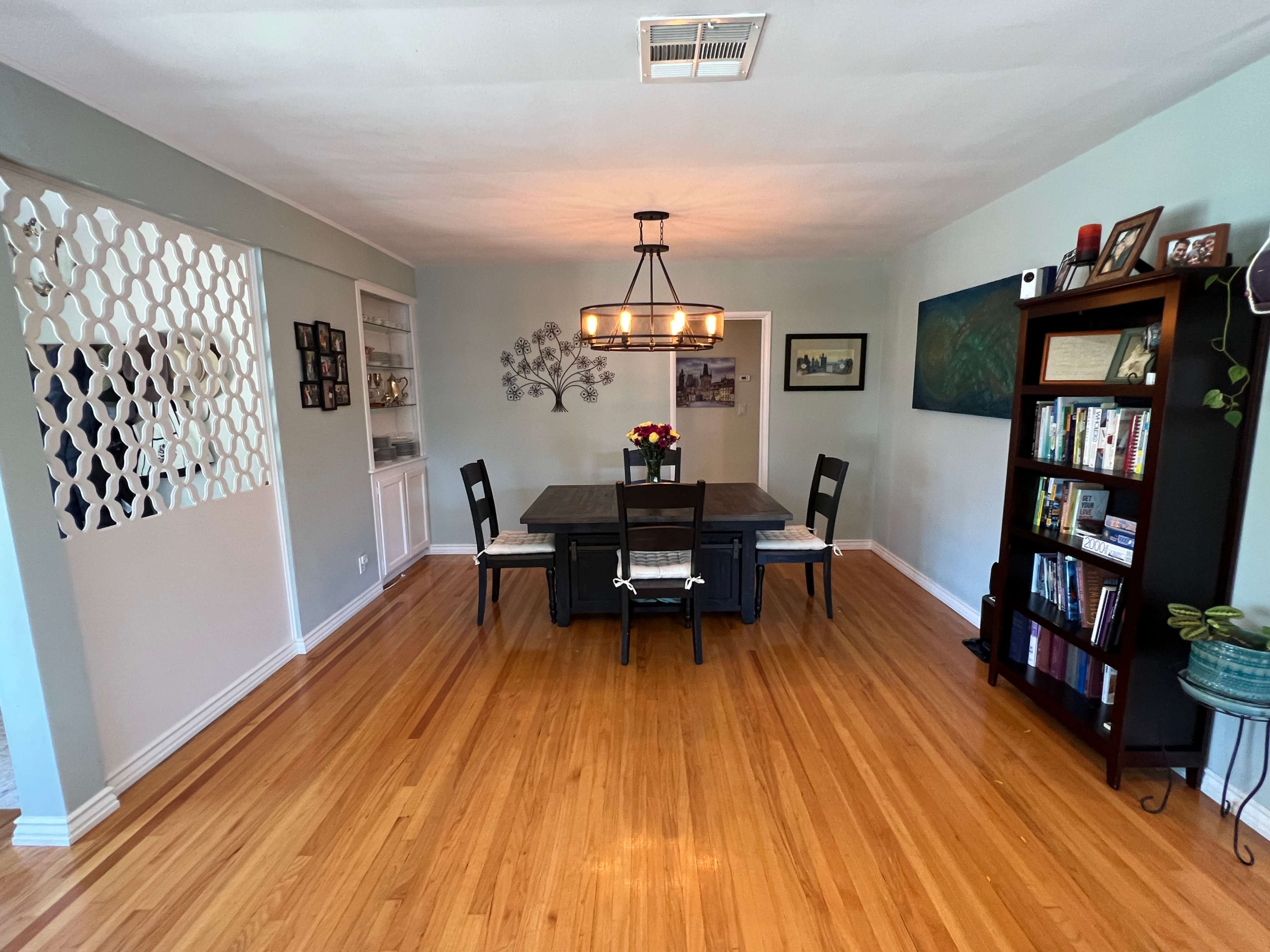 A spacious dining room featuring a large rectangular table surrounded by six chairs, with a decorative chandelier overhead and bookshelves along one wall.