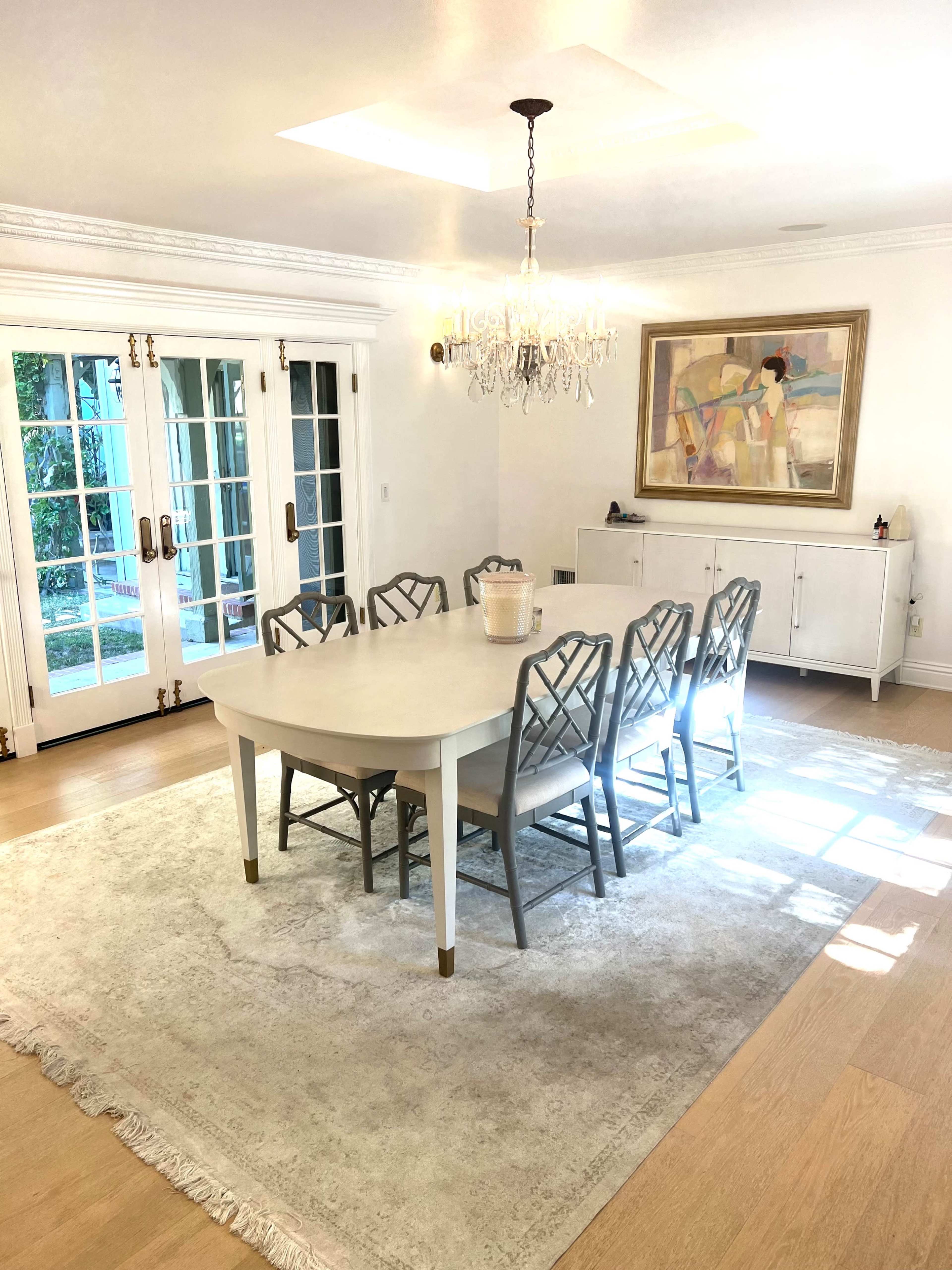 A dining room features a large table surrounded by six chairs, near a rug, a chandelier, and a sideboard, with glass doors leading to an outdoor area.