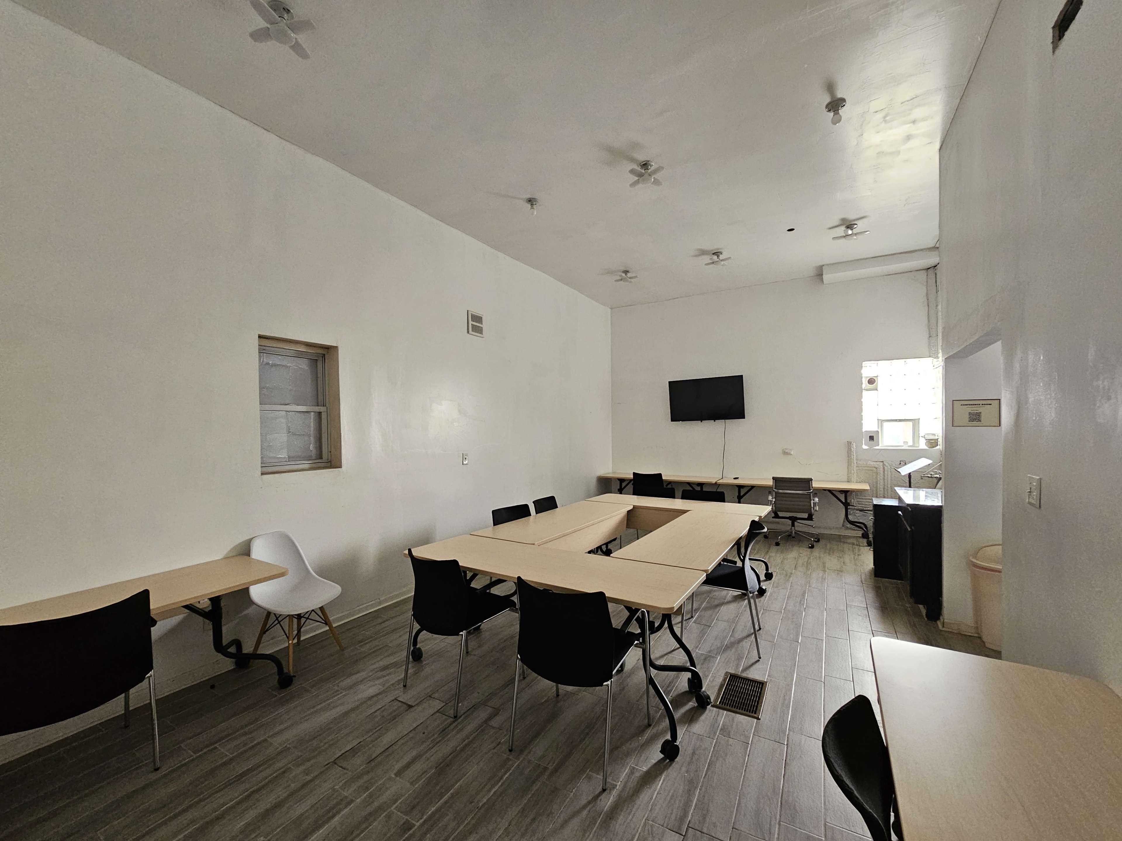 The image shows a sparsely furnished meeting room with several tables and chairs arranged, a wall-mounted TV, and minimal decor along the white walls.