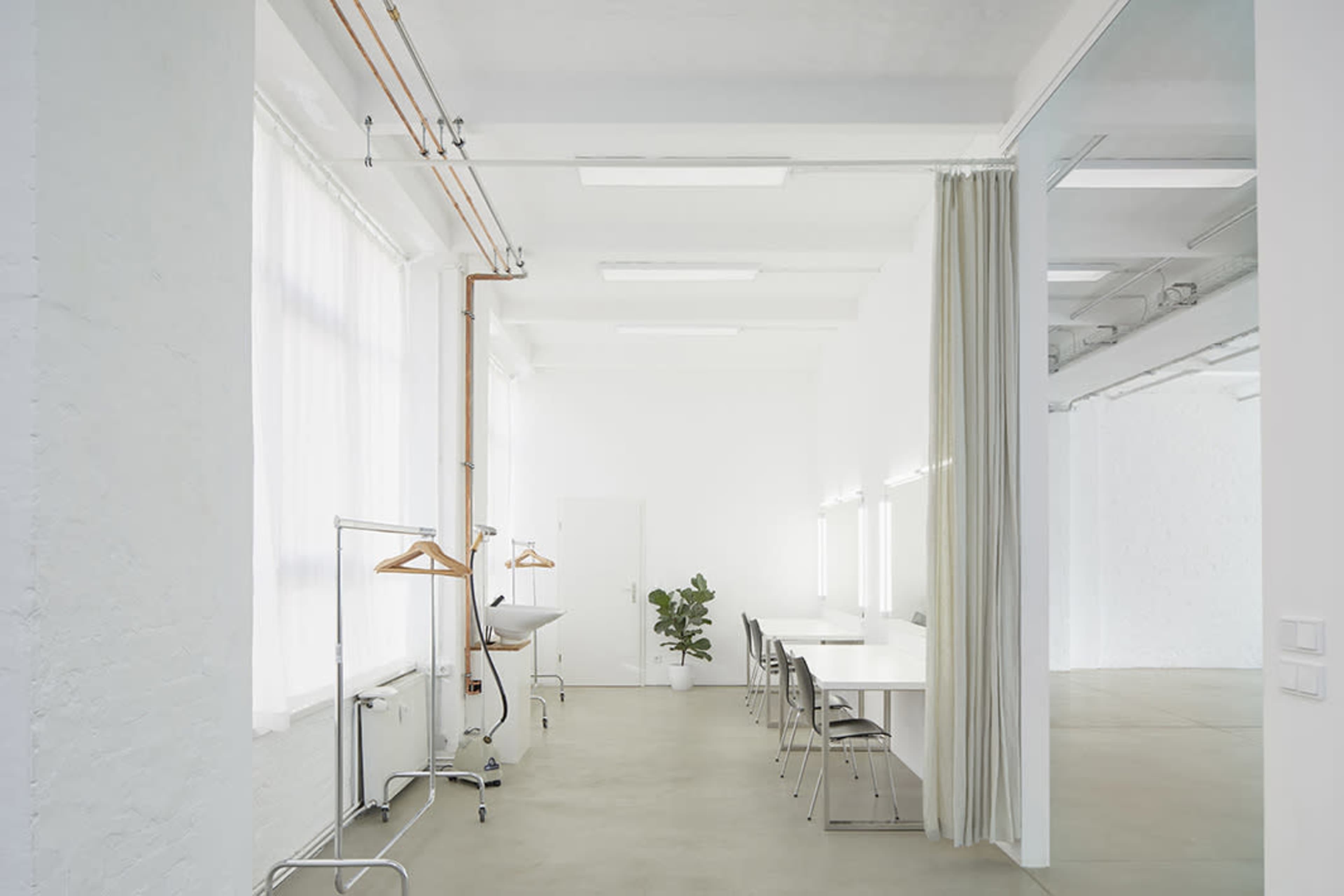 A bright, minimalistic room with white walls, a long table, chairs, and a small potted plant, along with clothing racks and large windows allowing natural light.