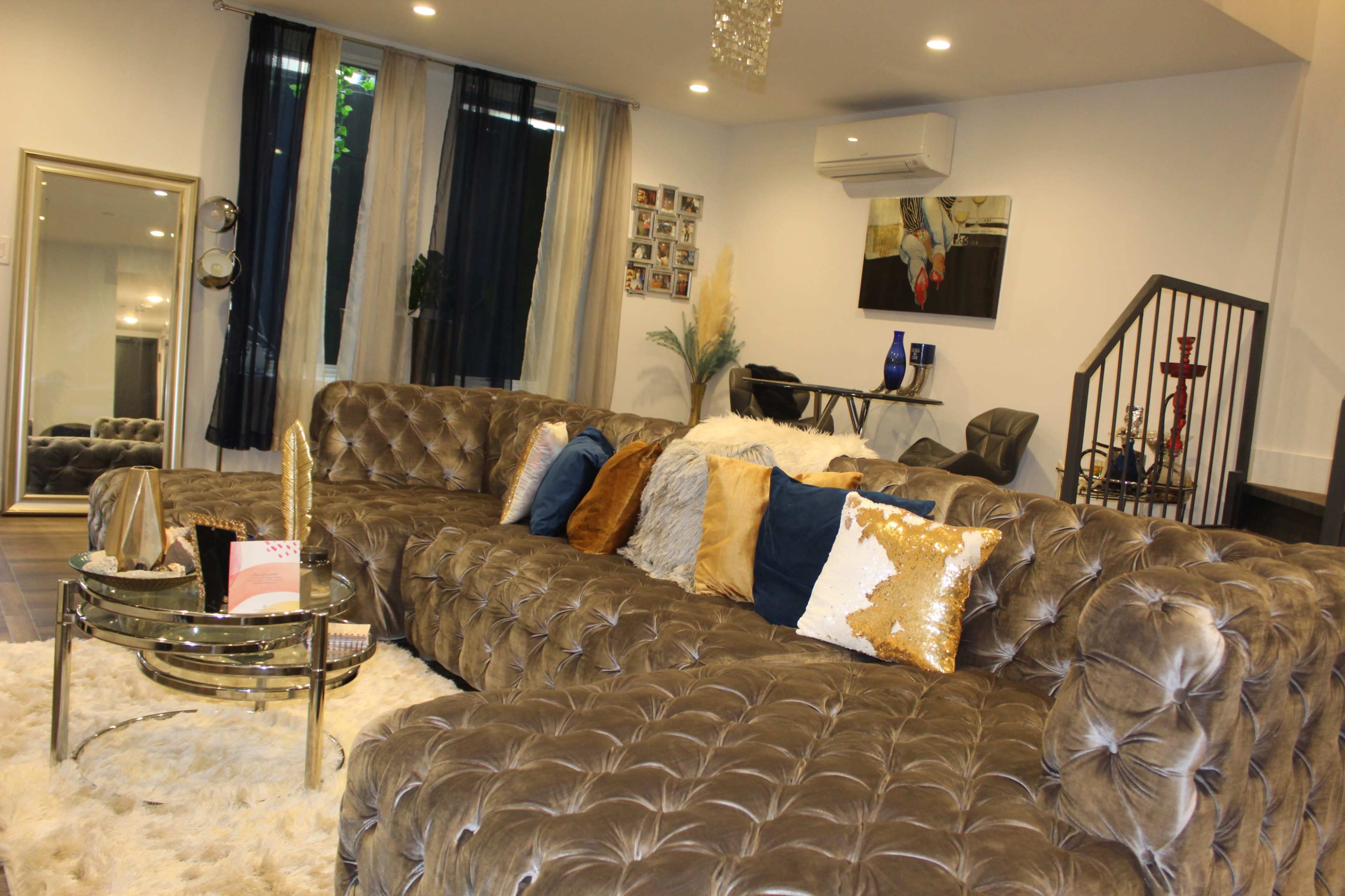 A modern living room featuring a large tufted gray sofa, decorative pillows, a glass coffee table, and a mix of light and dark drapes.