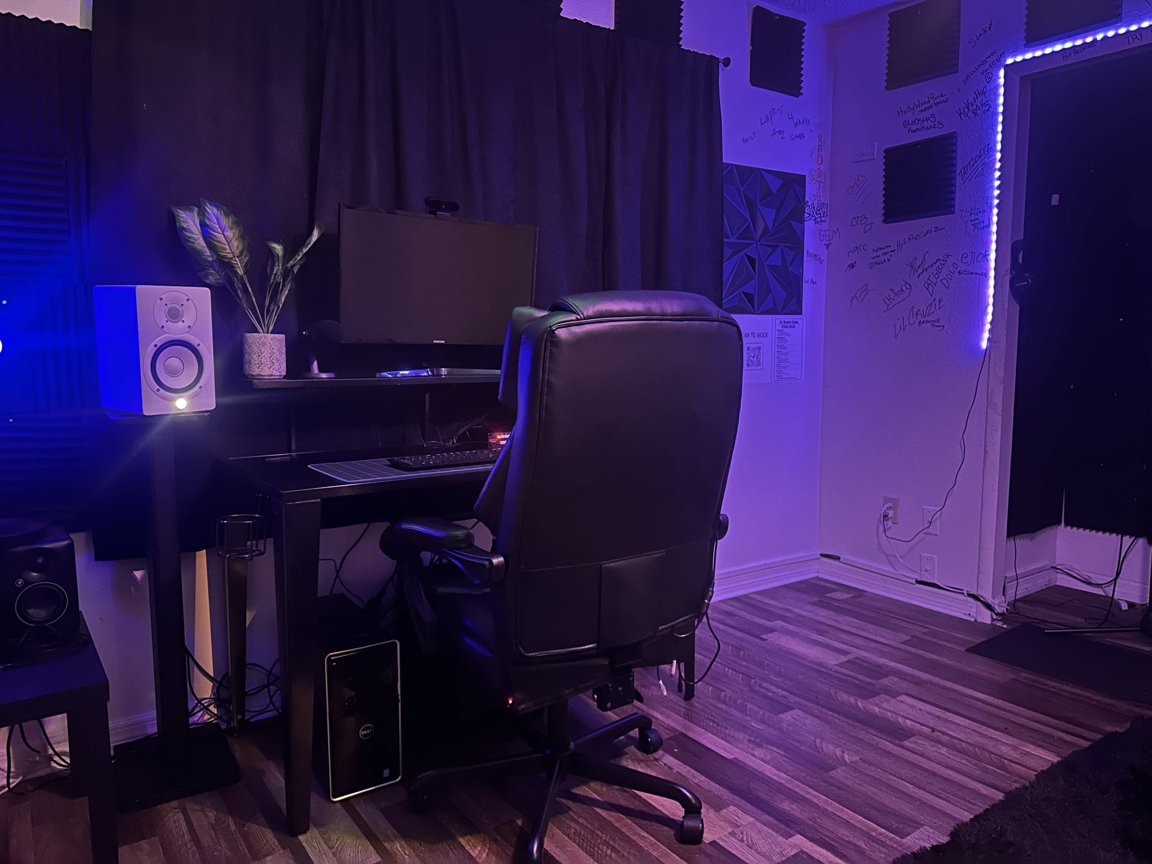 The image shows a workspace with a black desk, an office chair, a computer monitor, and a plant, set against a backdrop of dark curtains and decorative wall elements.