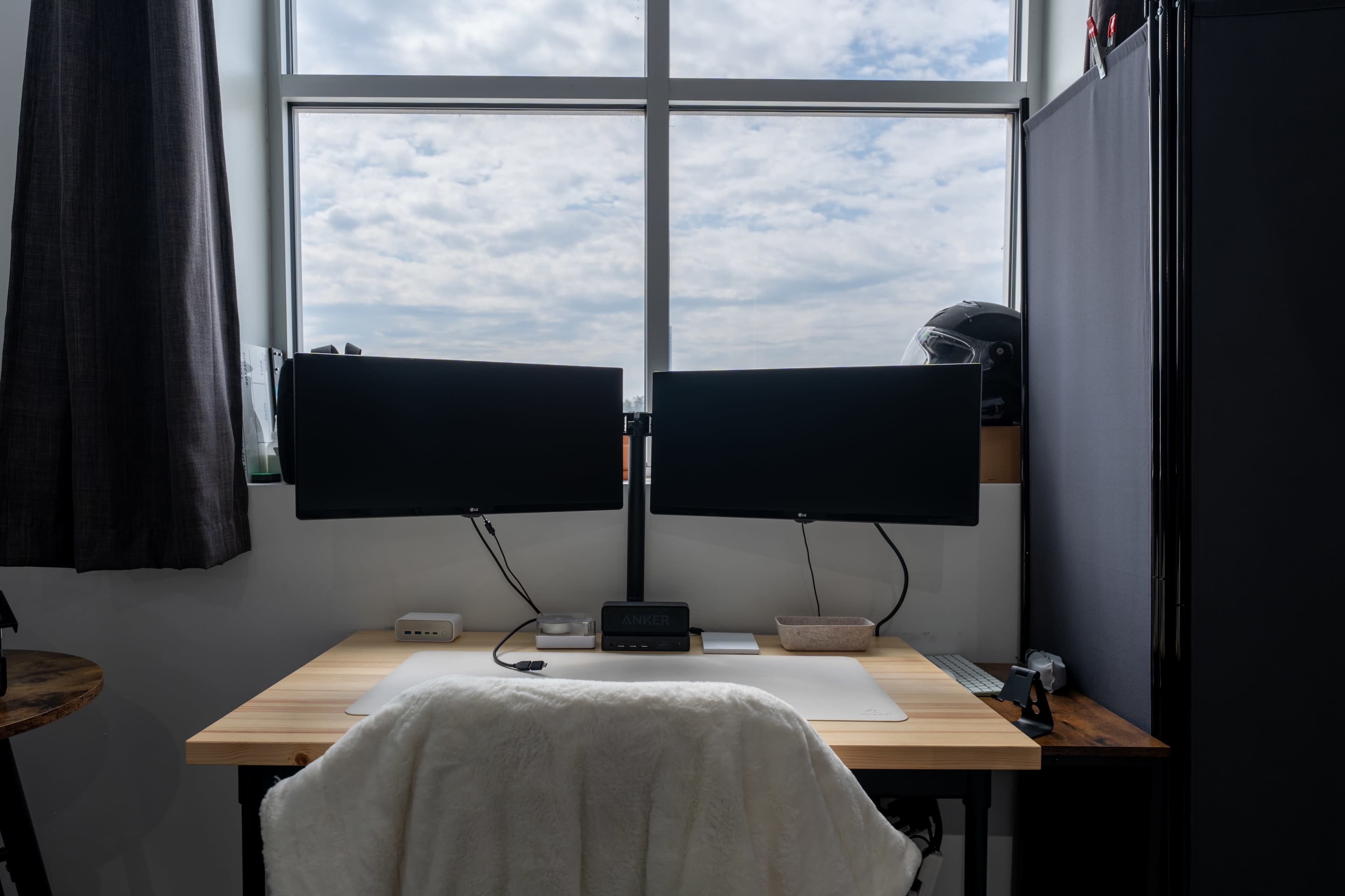 A desktop setup featuring two monitor screens, a light-colored desk with a keyboard and mouse, and a window with a cloudy sky in the background.