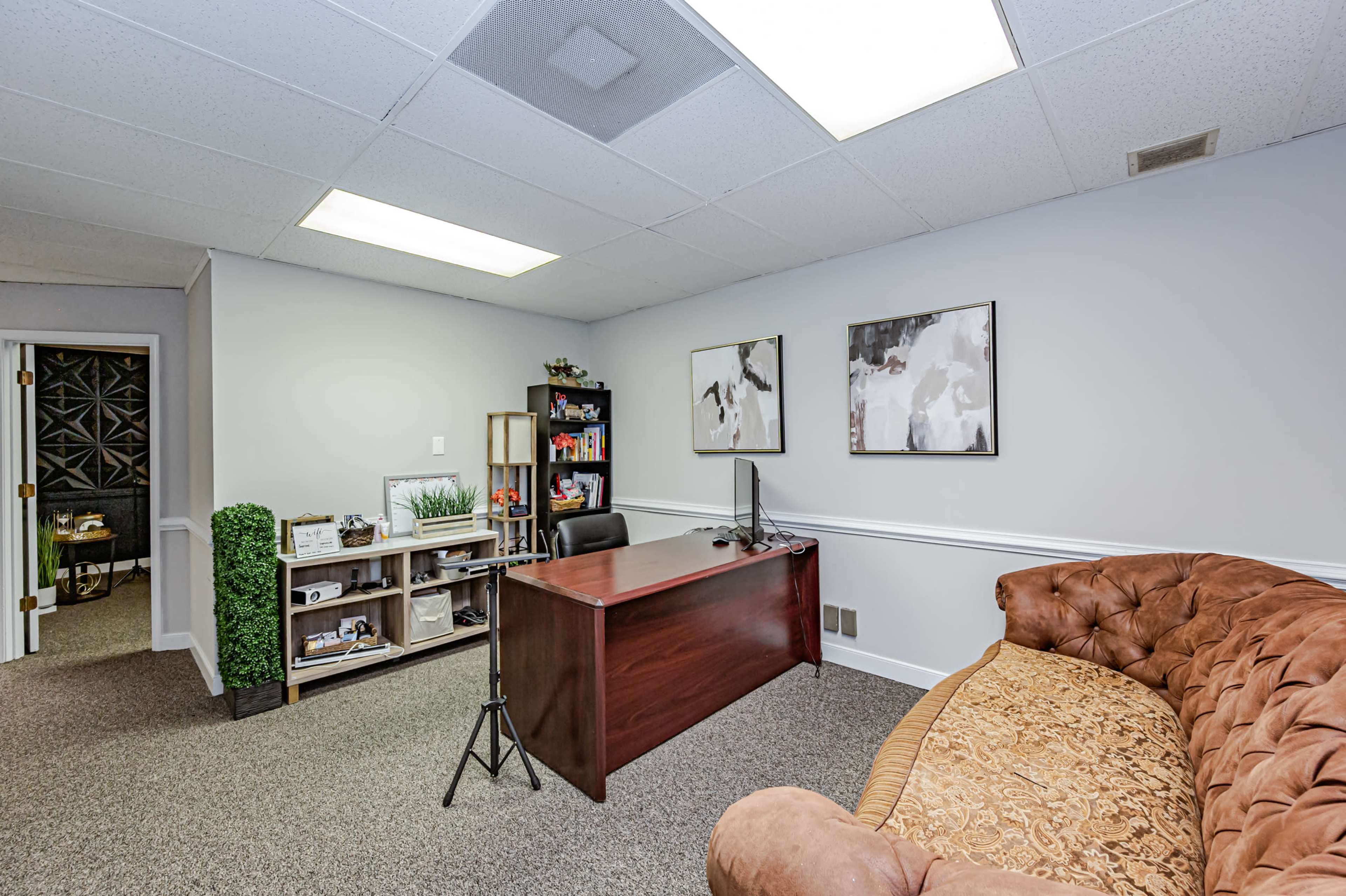 A small office space featuring a brown couch, a wooden desk with a computer monitor, and bookshelves filled with various items.