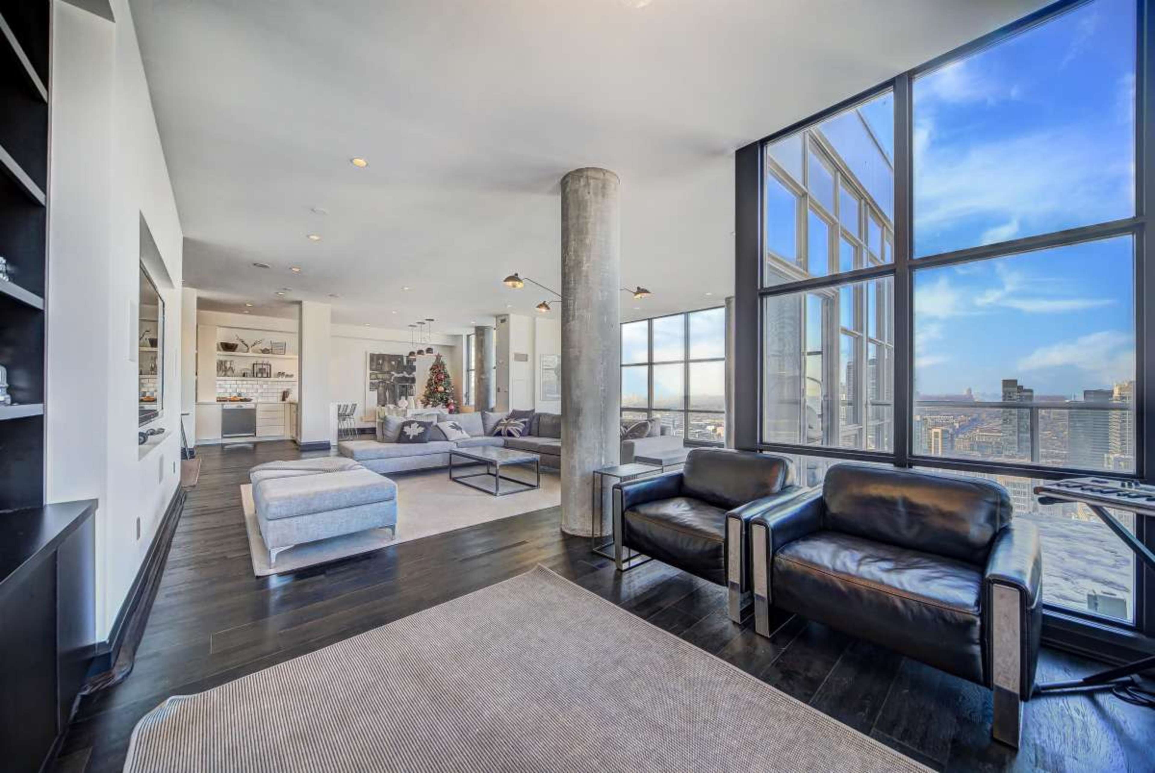 The image shows a modern, spacious living room with large windows, a sectional sofa, and black armchairs, overlooking a cityscape.