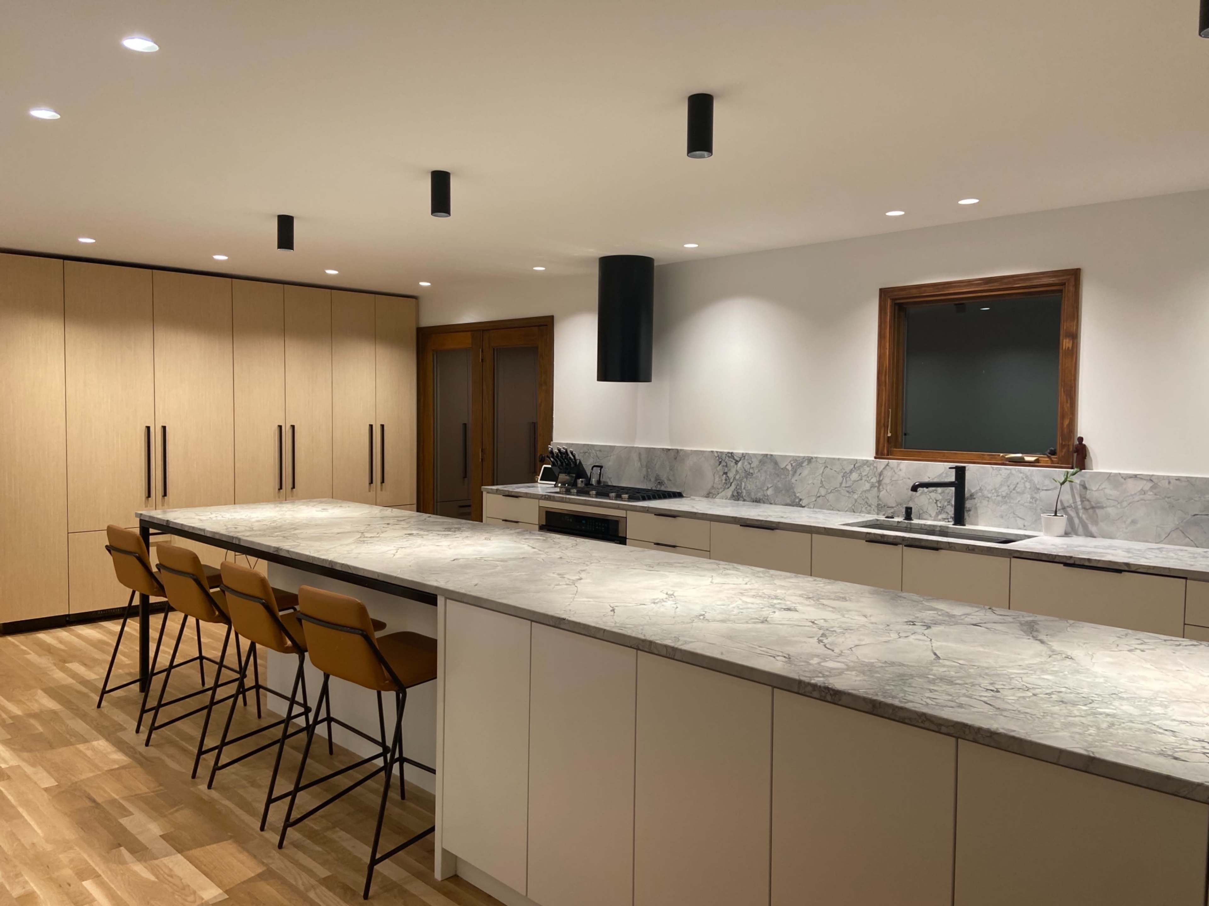 A modern kitchen features a large marble island with bar seating, wooden cabinetry, and a stove against a marble backsplash.