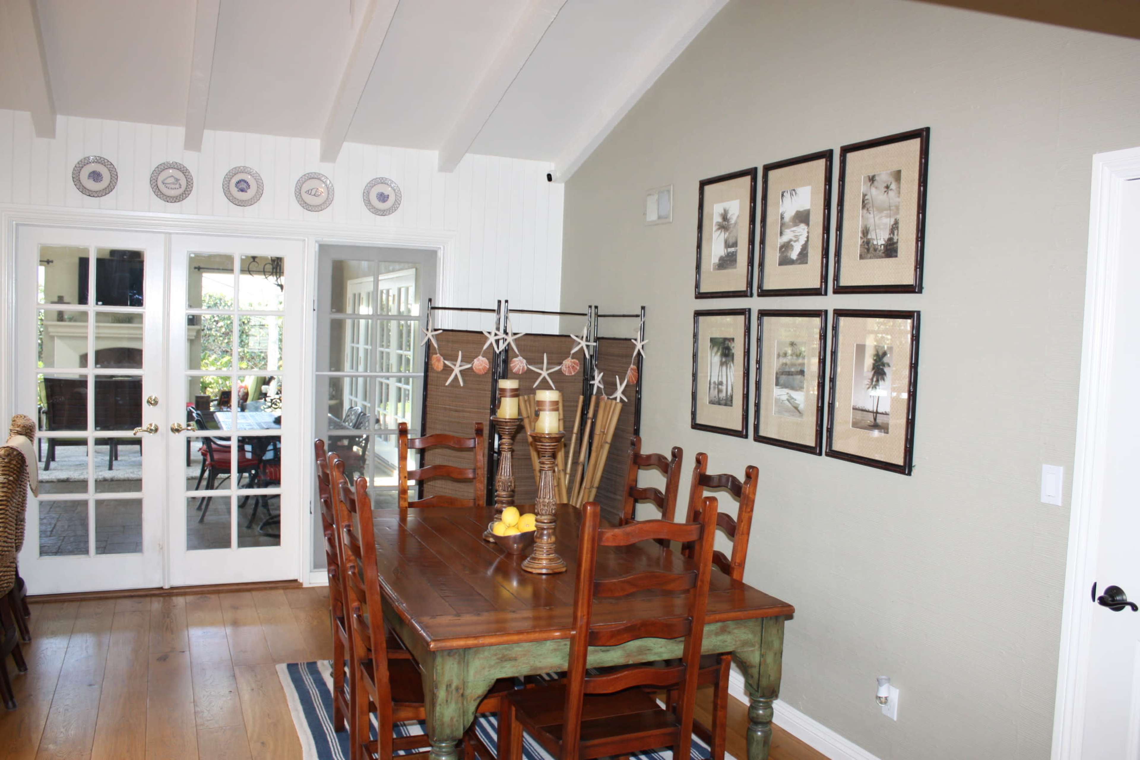 A wooden dining table with chairs sits beneath framed photographs on a wall, adjacent to a set of glass doors leading to an outdoor area.