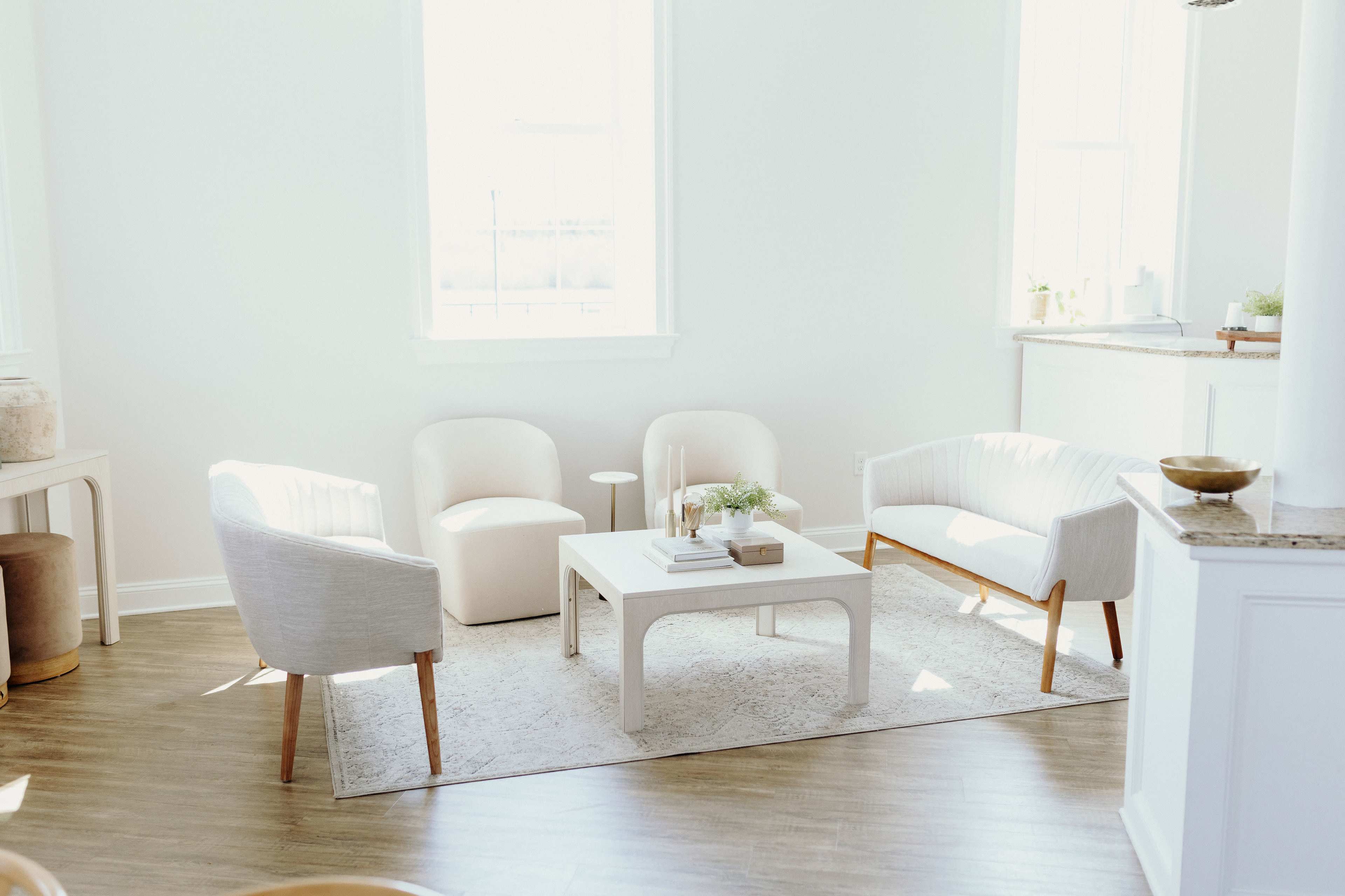 A bright, minimalistic living room with two white armchairs, one light-colored chair, and a square table on a light rug.