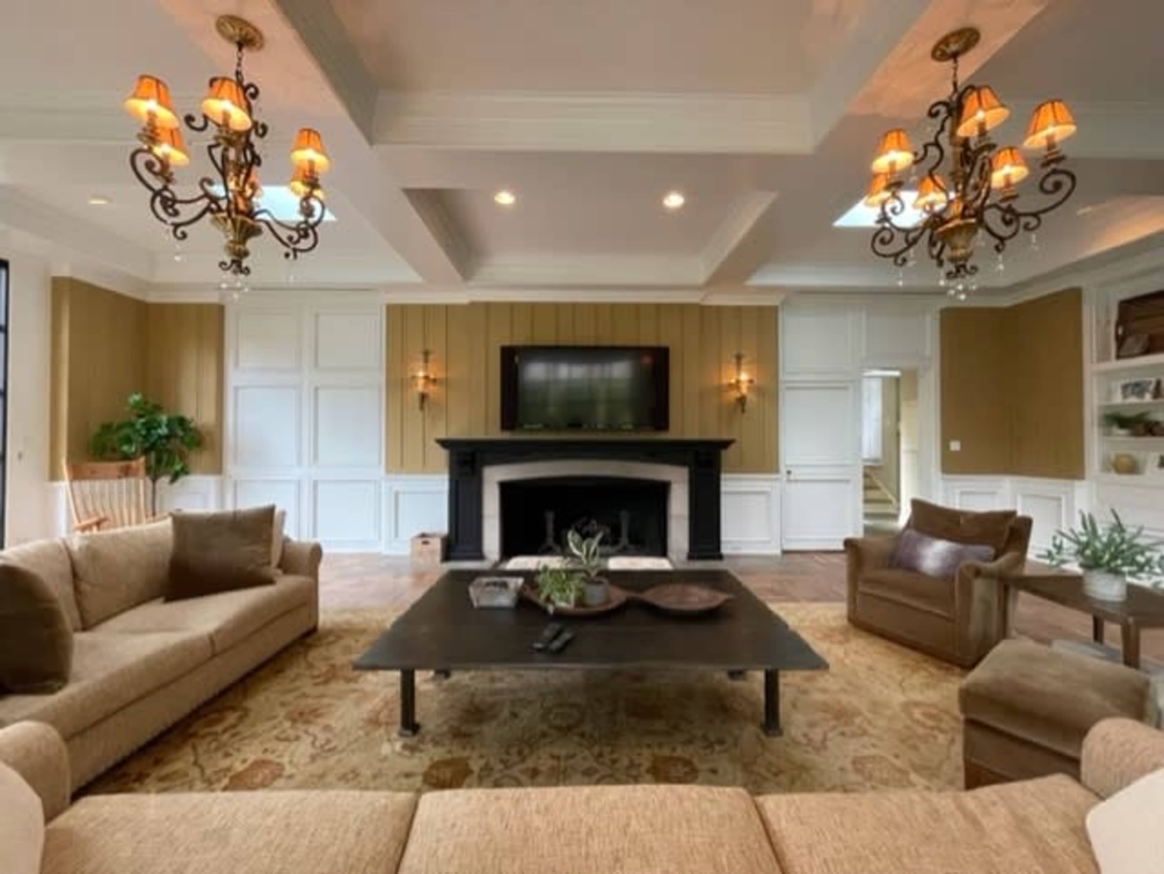 The living room features a central coffee table surrounded by beige sofas and a leather armchair, with a television mounted above a black fireplace and two chandeliers hanging from the ceiling.