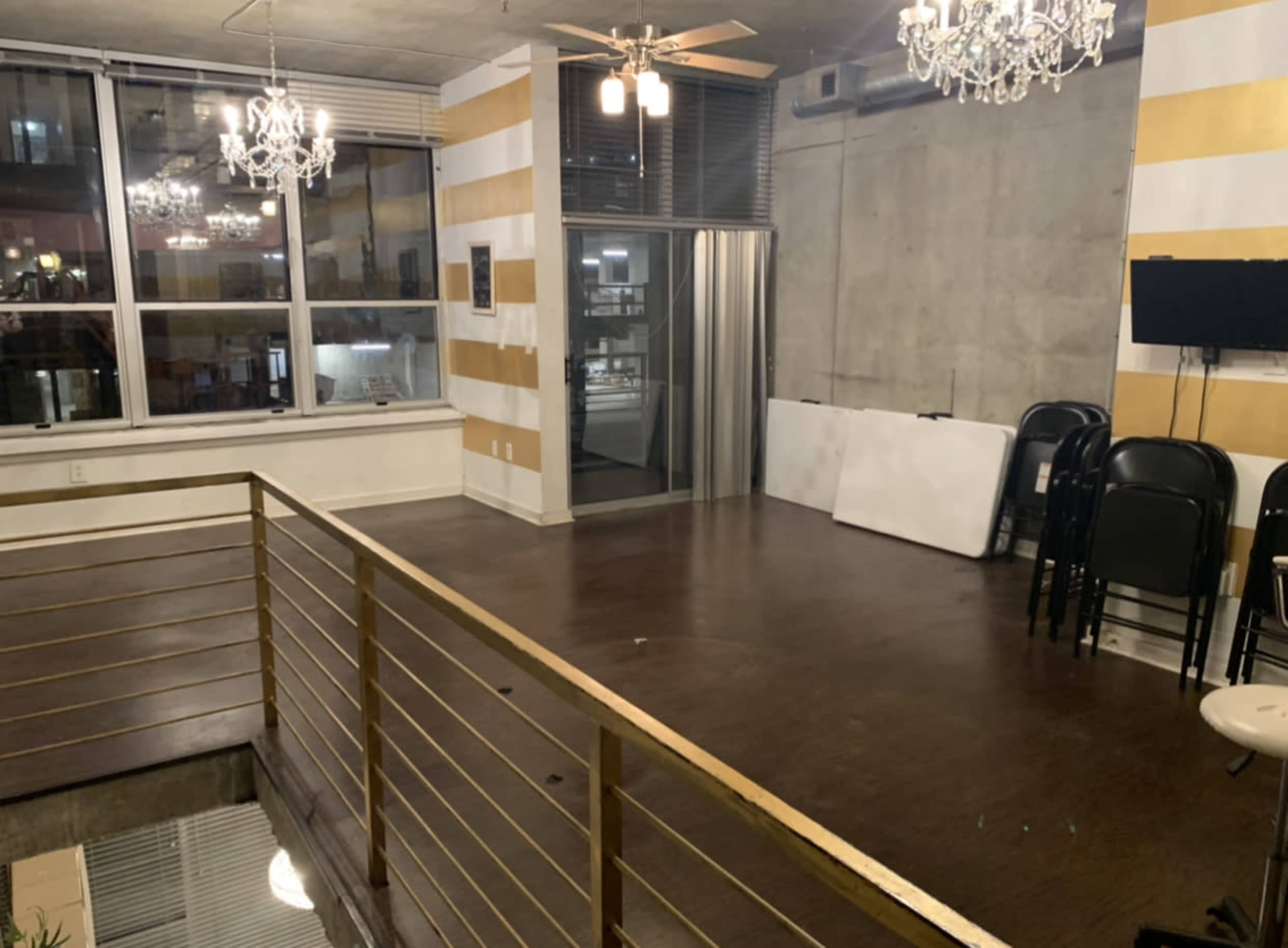 A spacious room with a wooden floor, large windows, and a railing overlooking a lower area, featuring decorative chandeliers and chairs stacked against a wall.