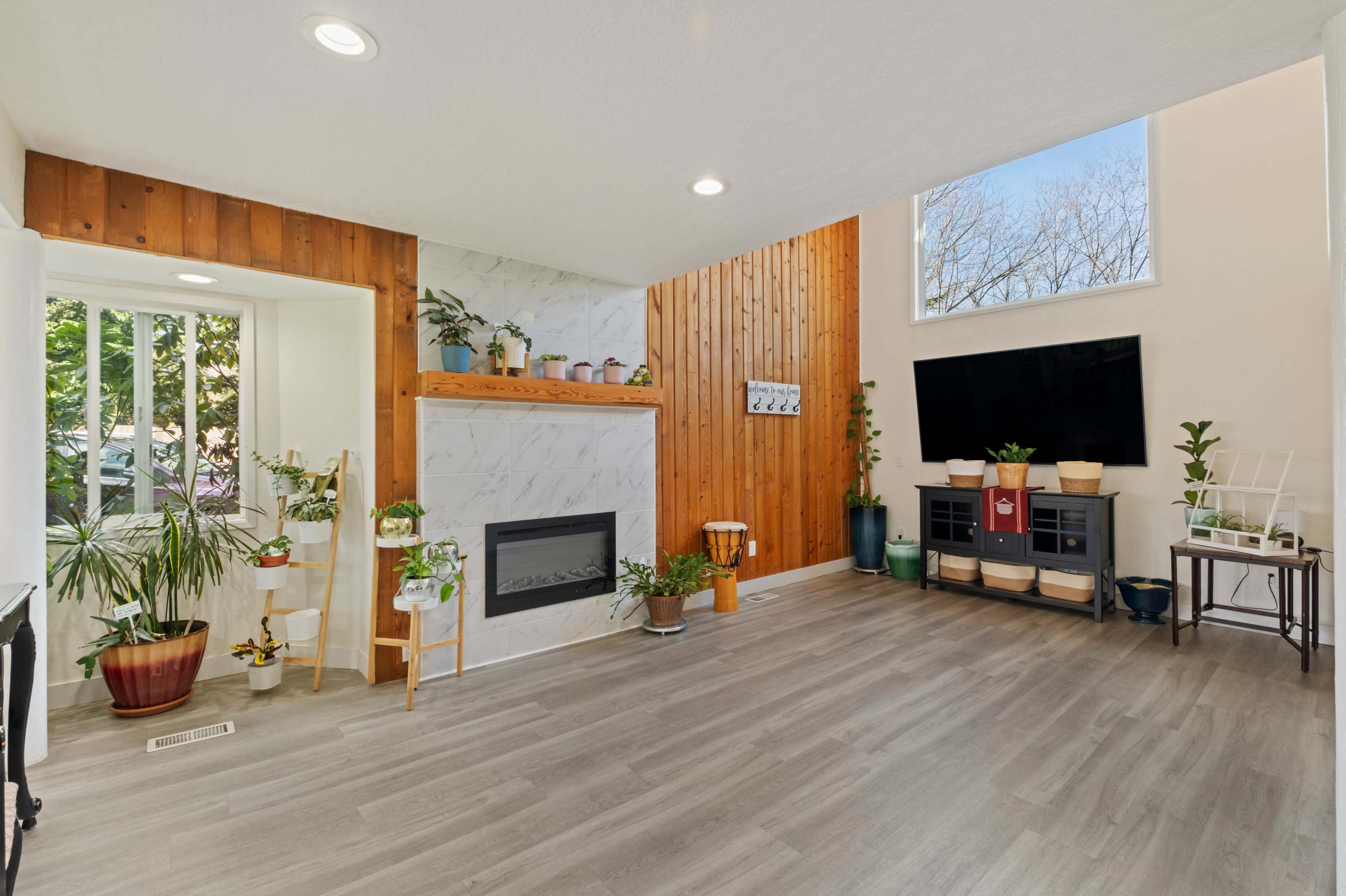 The image shows a bright living room featuring a white marble fireplace, wooden accent walls, and various potted plants arranged throughout the space.