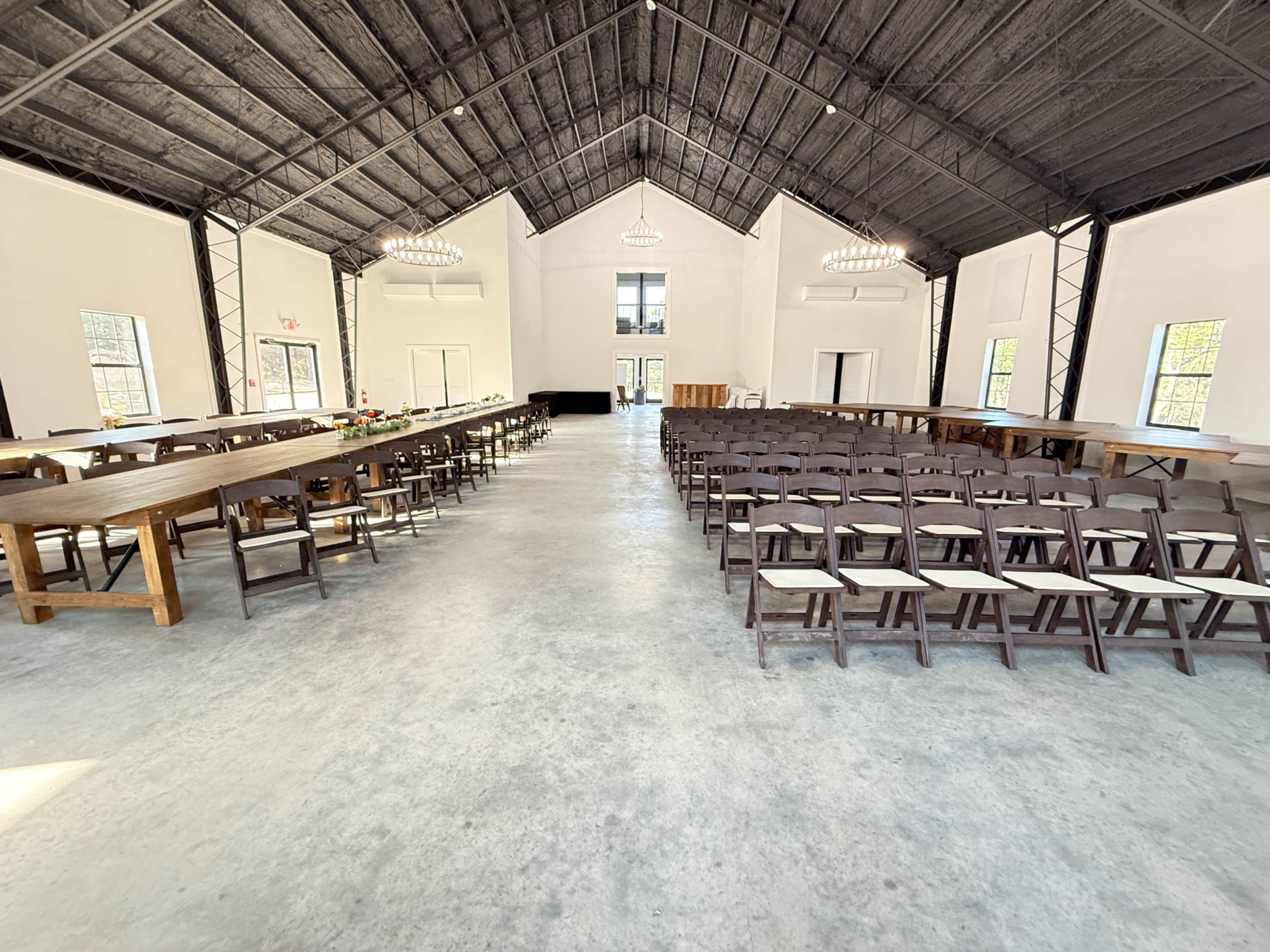 The image shows a spacious event hall with a high ceiling, featuring rows of wooden chairs facing a central stage area, flanked by long dining tables on either side.
