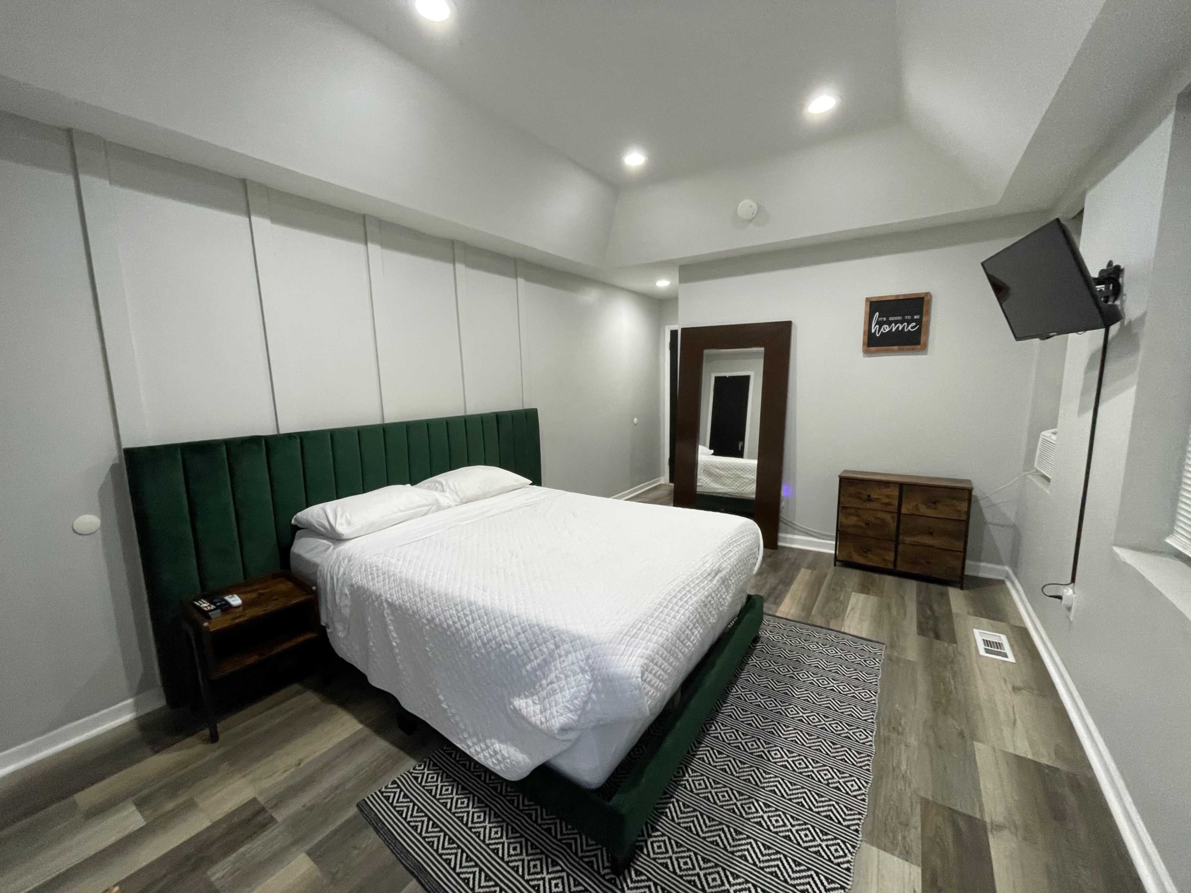 The image shows a modern bedroom with a green upholstered bed, a wooden nightstand and dresser, a flat-screen TV mounted on the wall, and a large mirror reflecting the room.