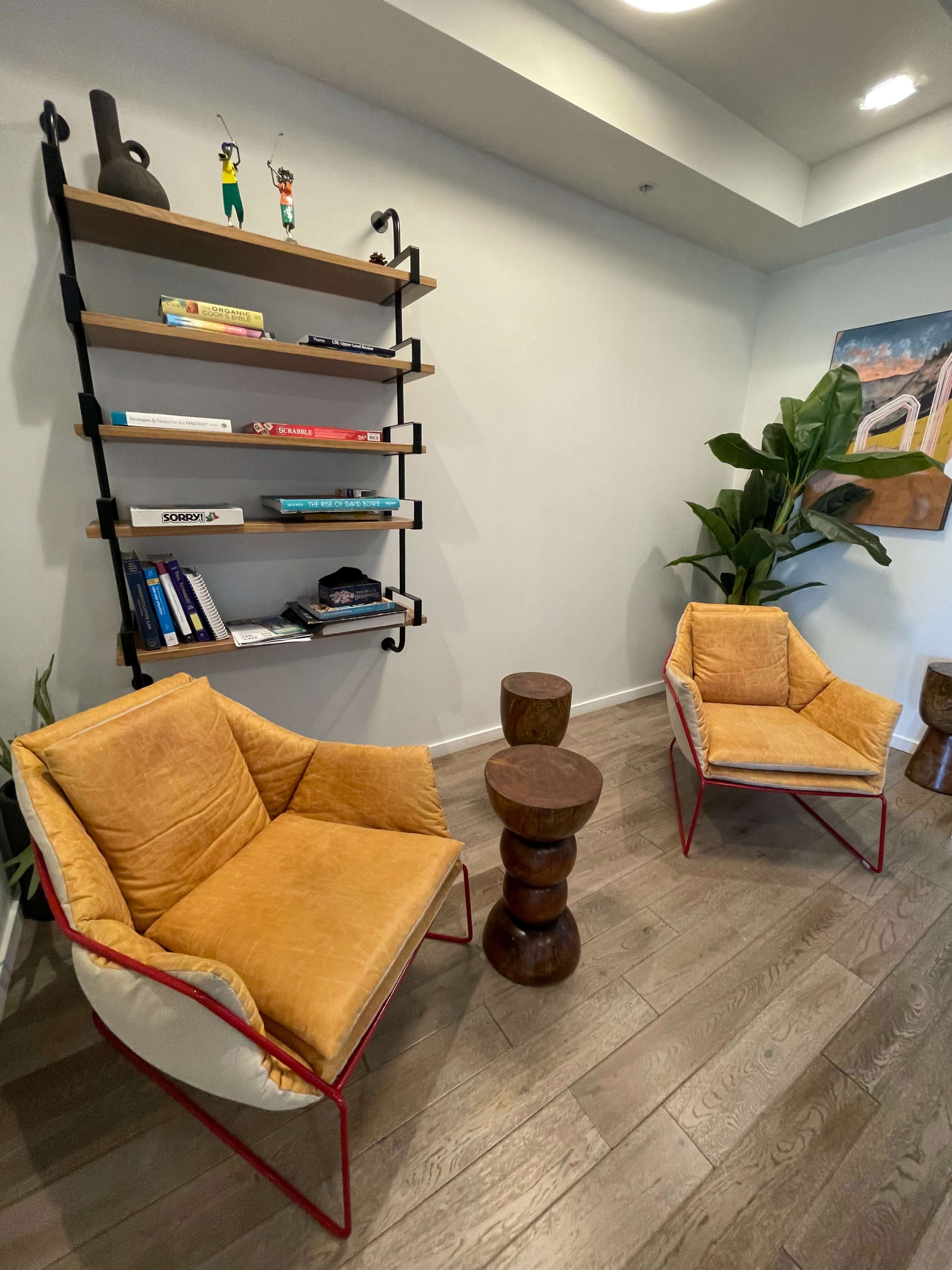 The image shows a modern interior featuring two yellow chairs, a wooden side table, and a wall shelf with books and decorations.