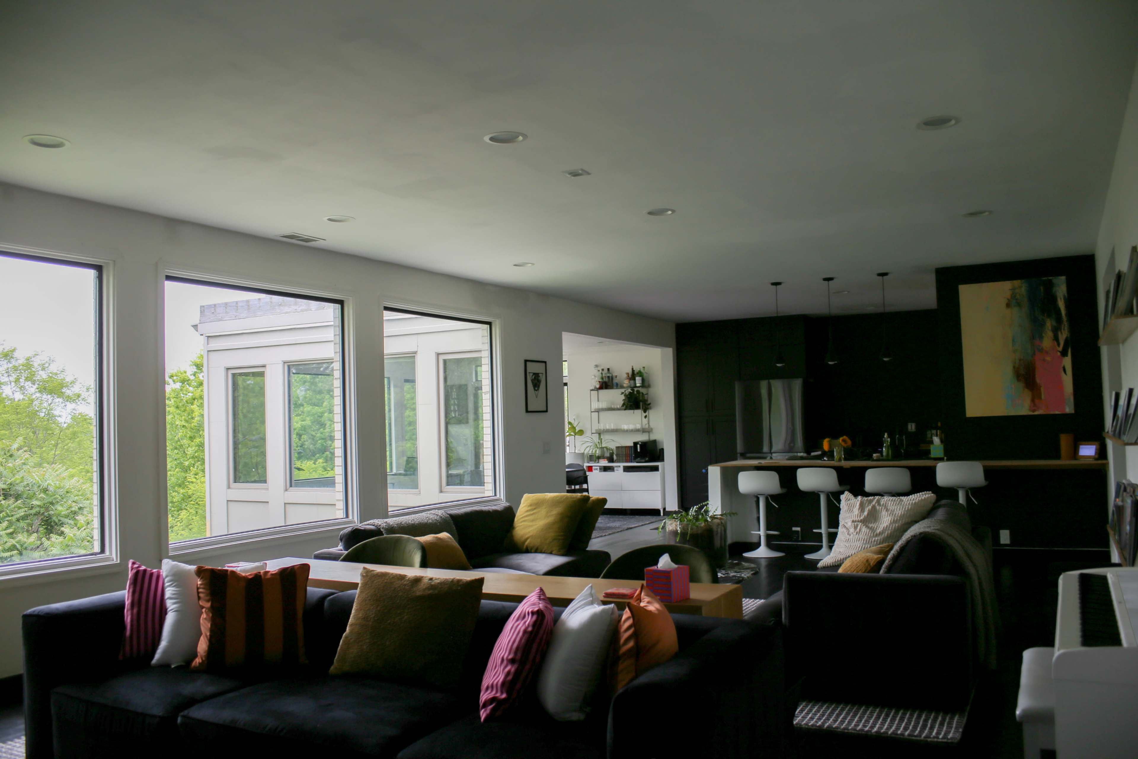 The image depicts a modern living room with large windows, a sectional sofa adorned with pillows, and an adjacent kitchen area featuring bar stools.