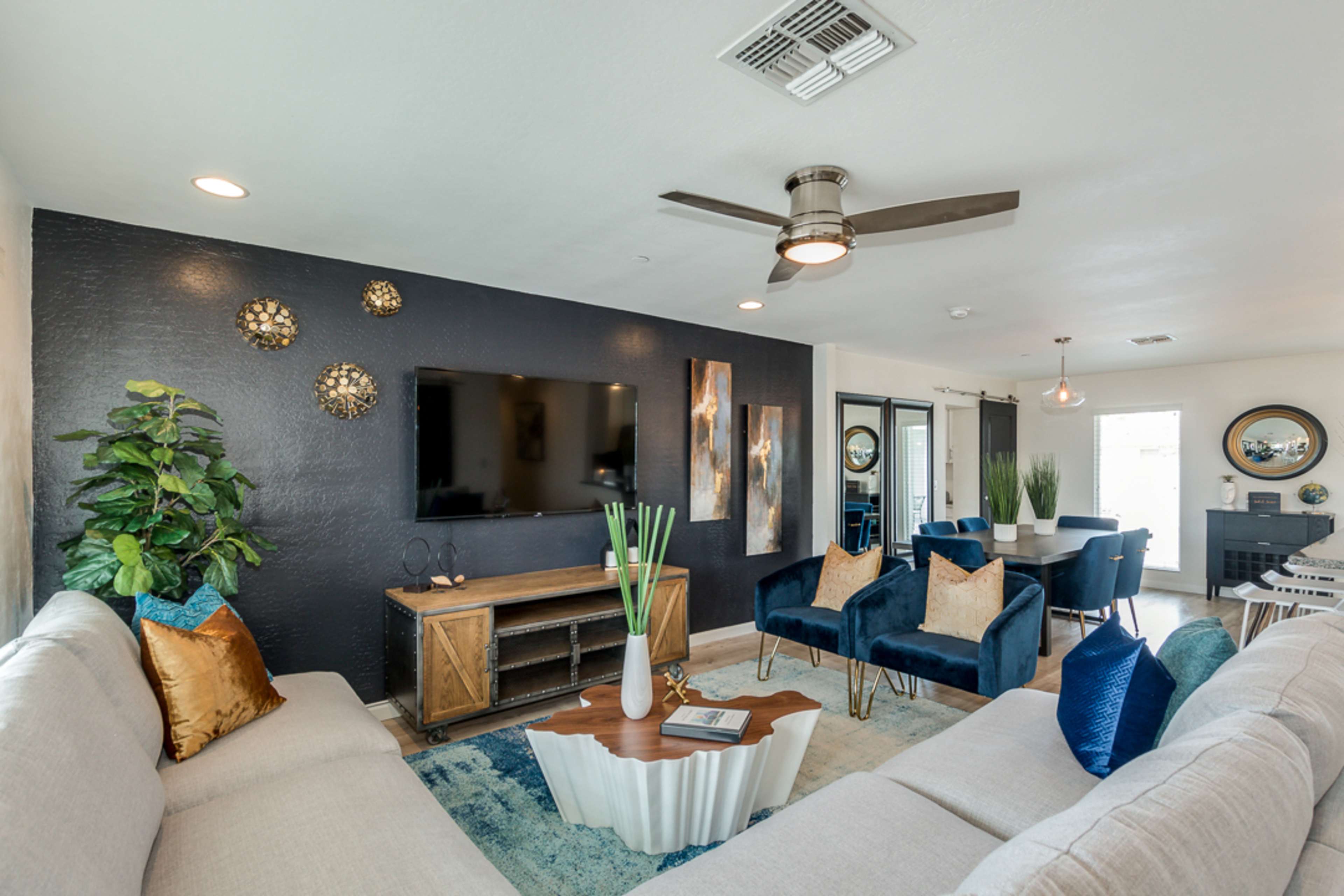 The living room features a dark accent wall with modern artwork, a mounted television, a wooden coffee table, and two blue armchairs, surrounded by neutral-toned sofas and greenery.