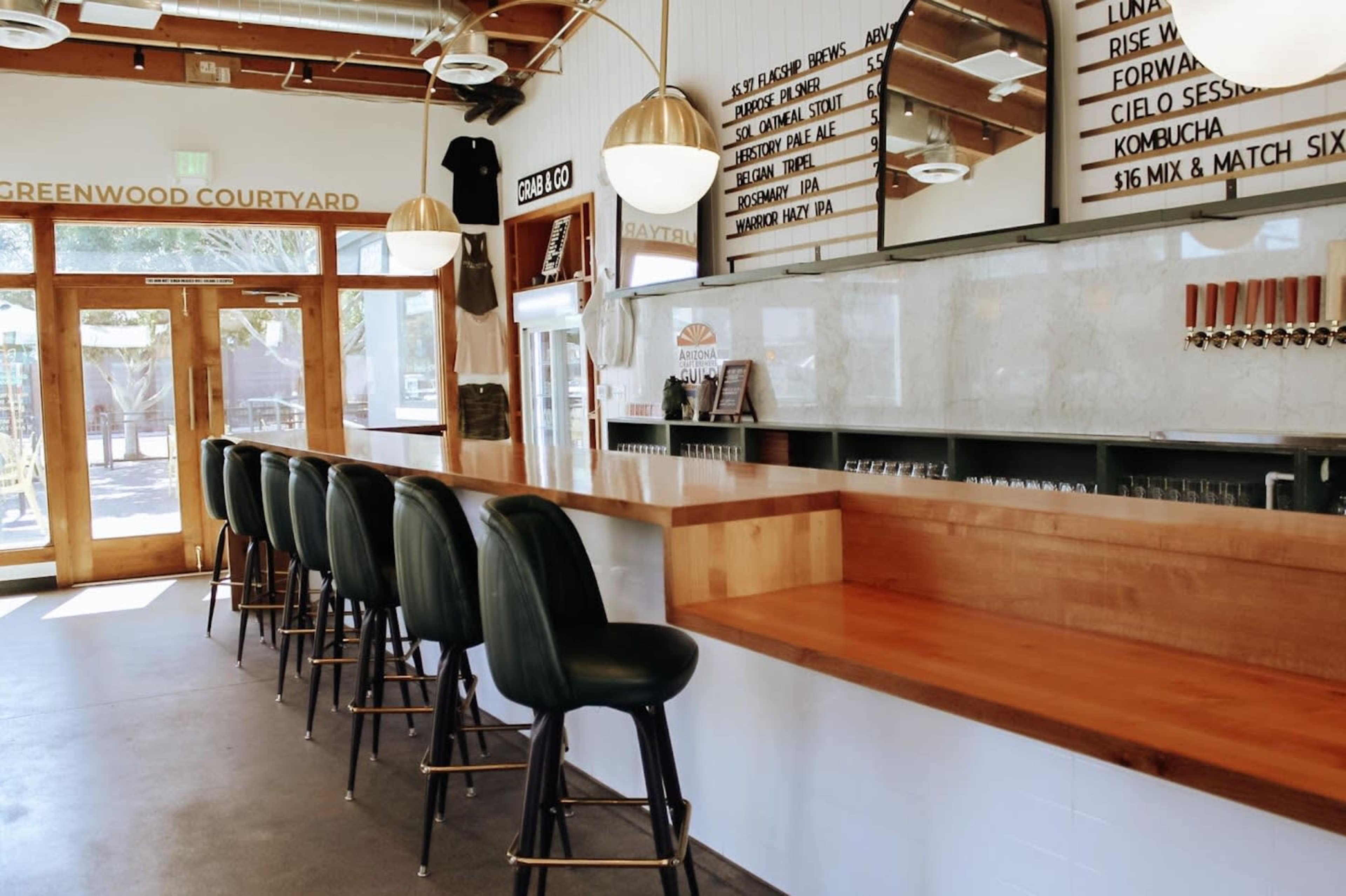 The interior of a cafe features a long wooden bar with green leather stools, large windows allowing natural light, and a menu displayed on the wall.