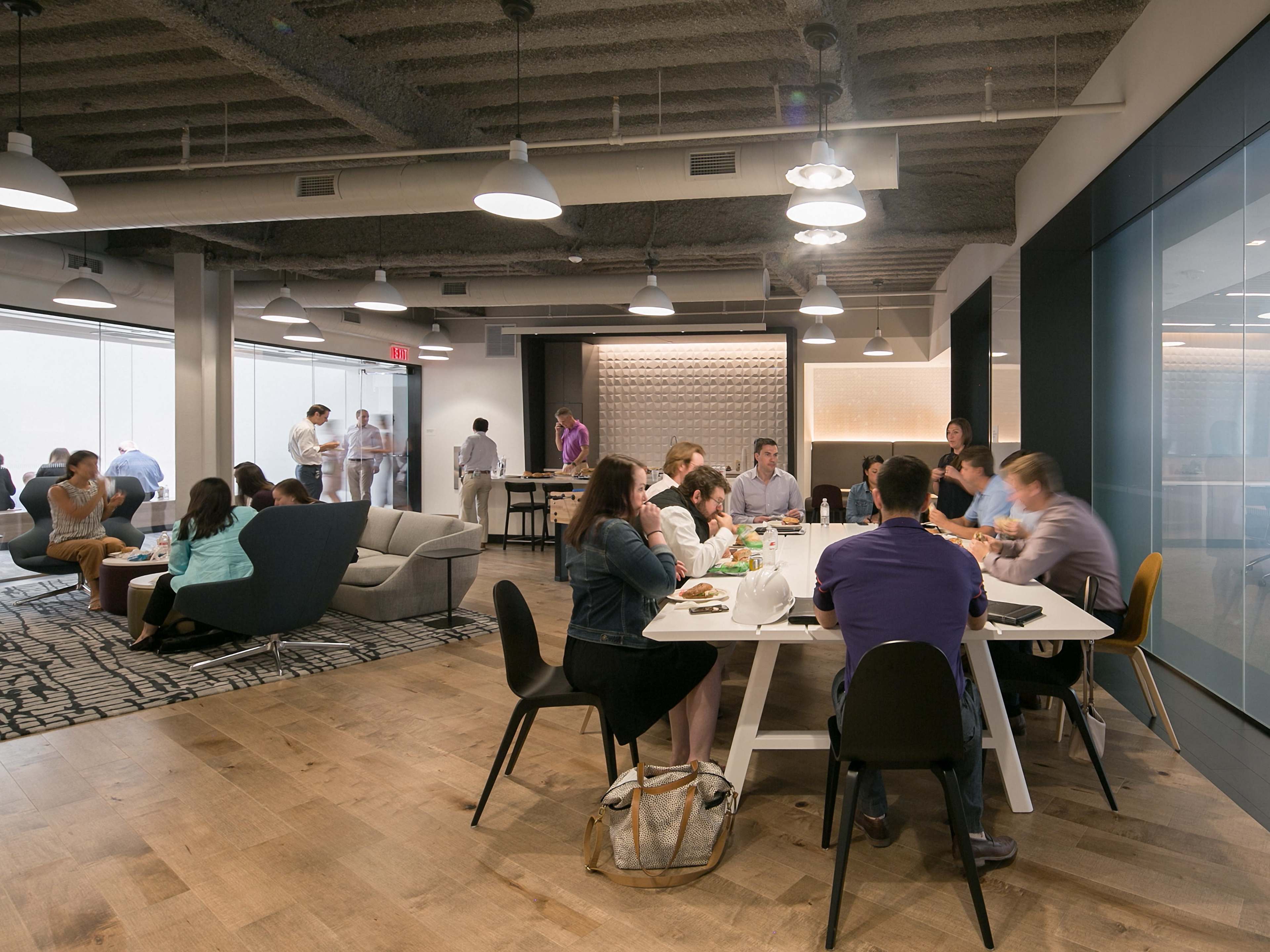 A group of people is gathered in a modern office space, participating in discussions and working at tables while others mingle in the background.