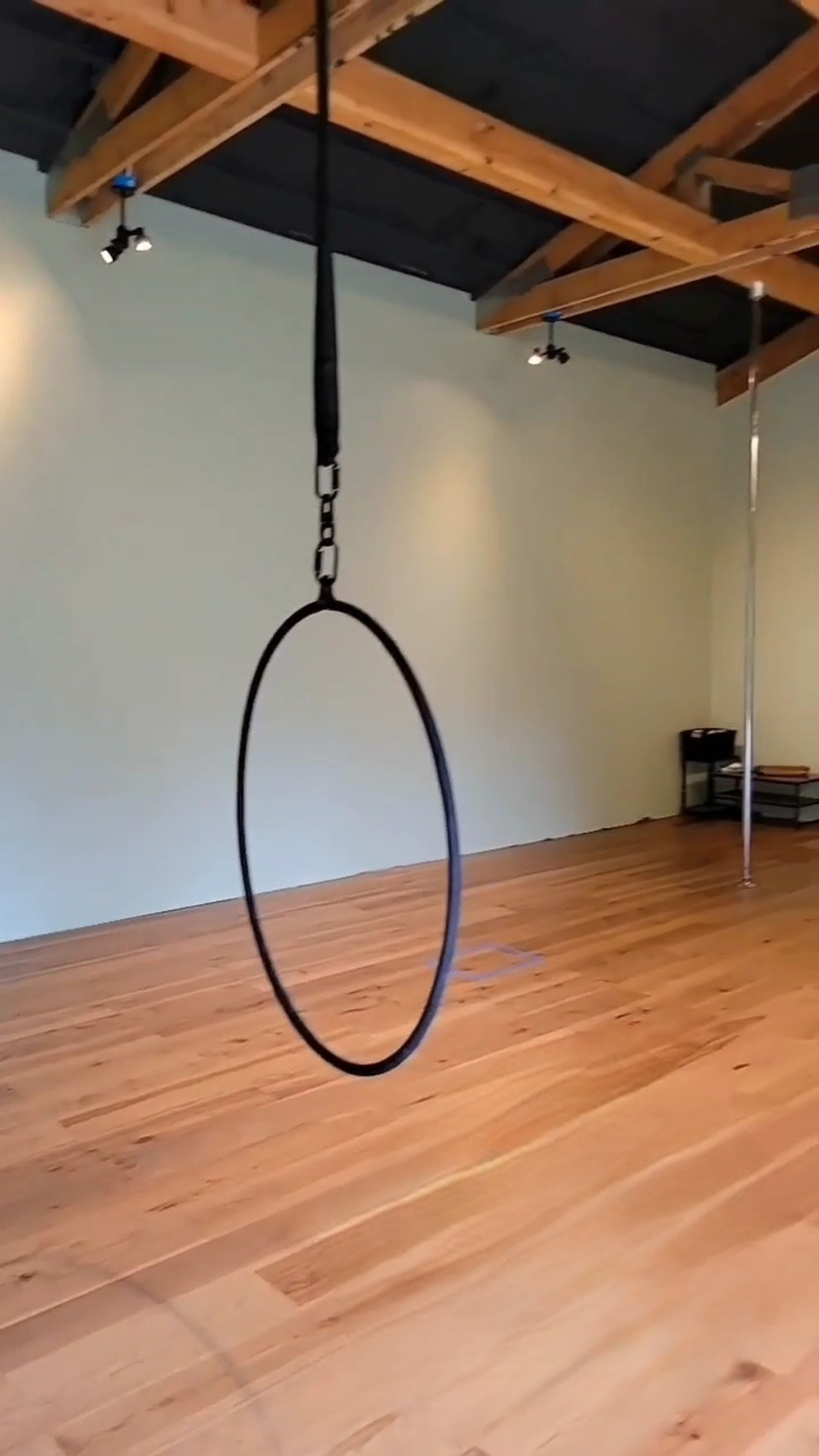 The image shows a spacious room with wooden floors and a suspended black circular hoop hanging from the ceiling.