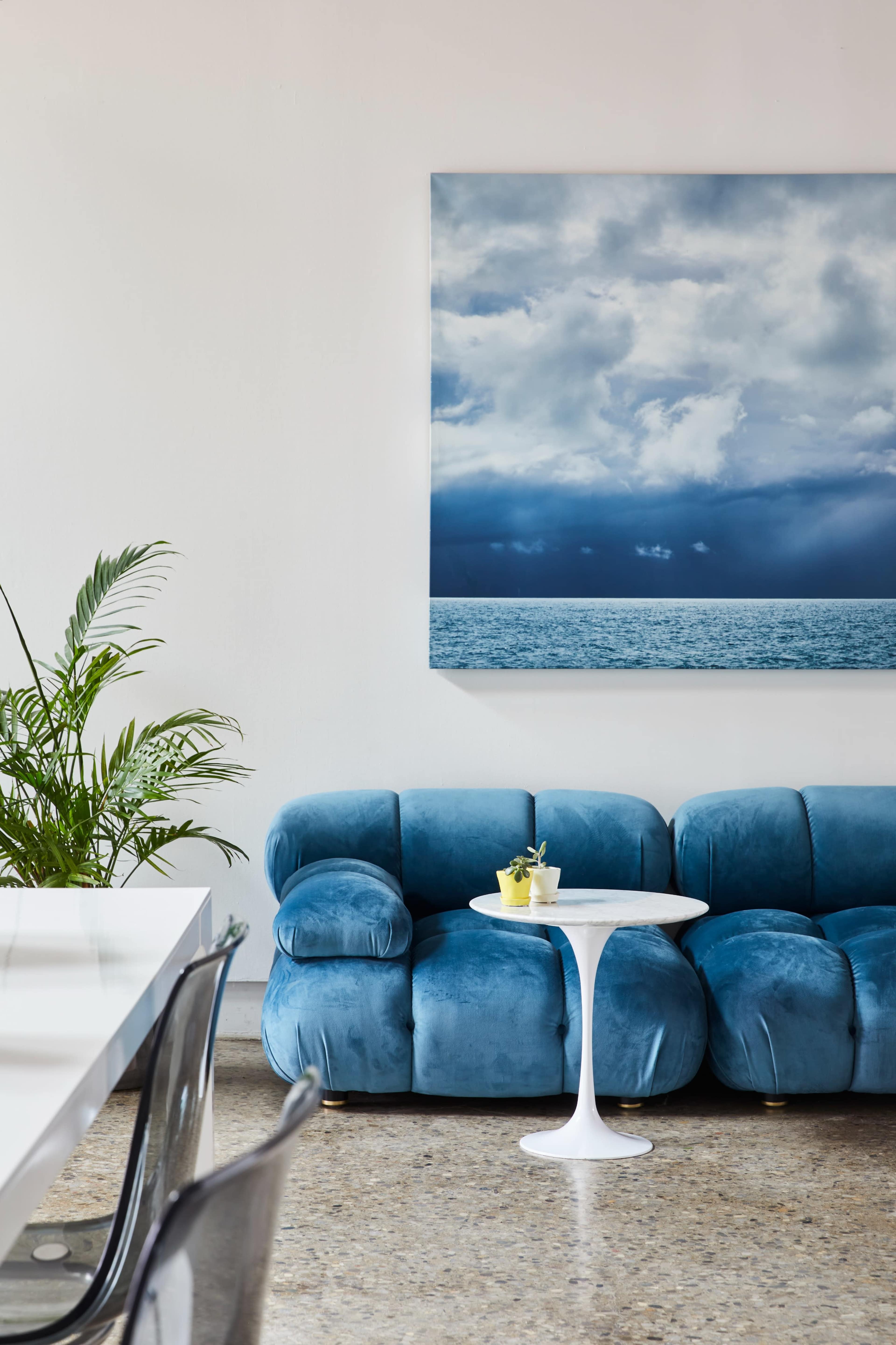 A blue velvet sofa with a white coffee table and a small plant is arranged in a modern interior featuring a large painting of clouds and ocean.
