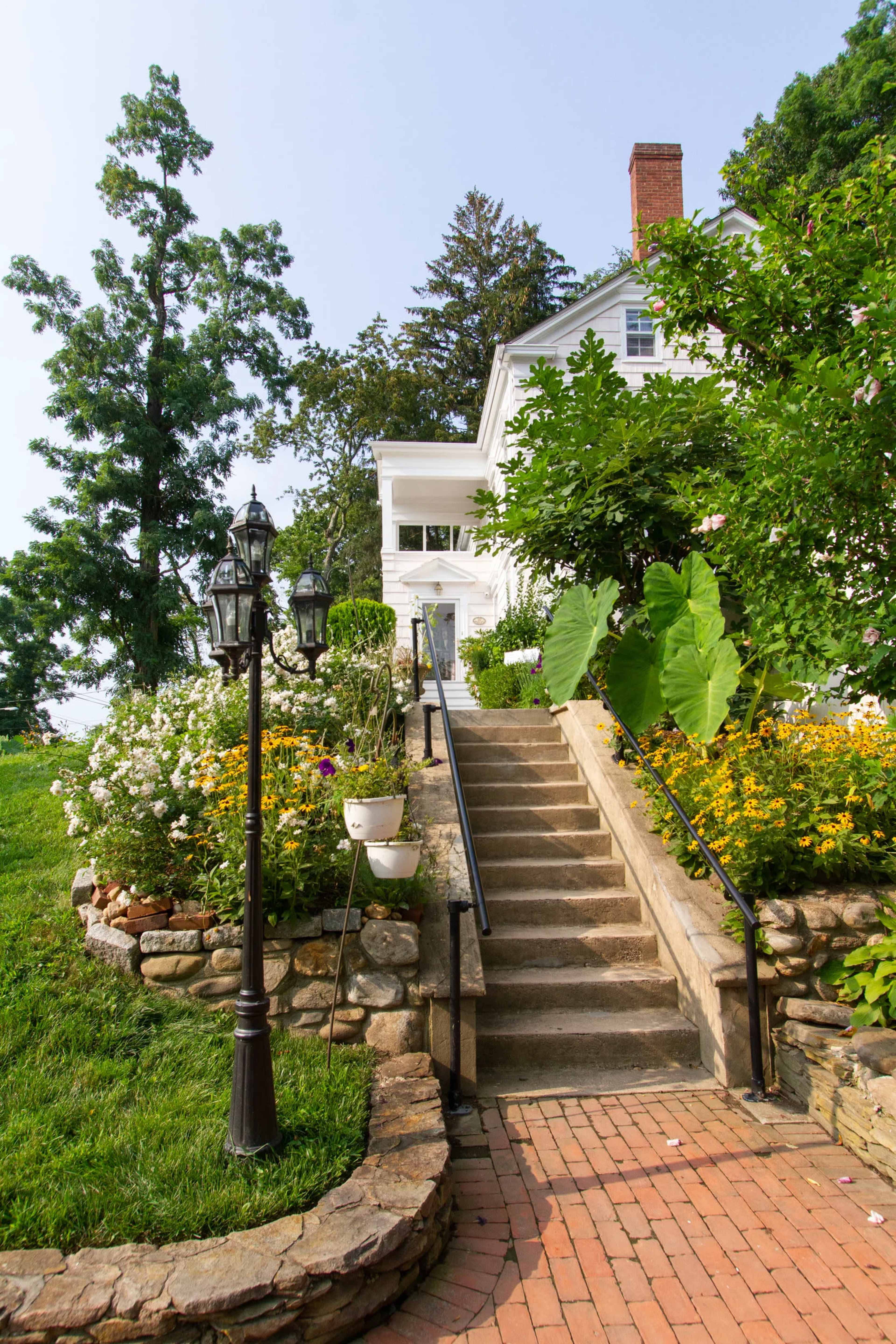 A stone pathway leads to a white house, flanked by lush greenery and colorful flower beds.