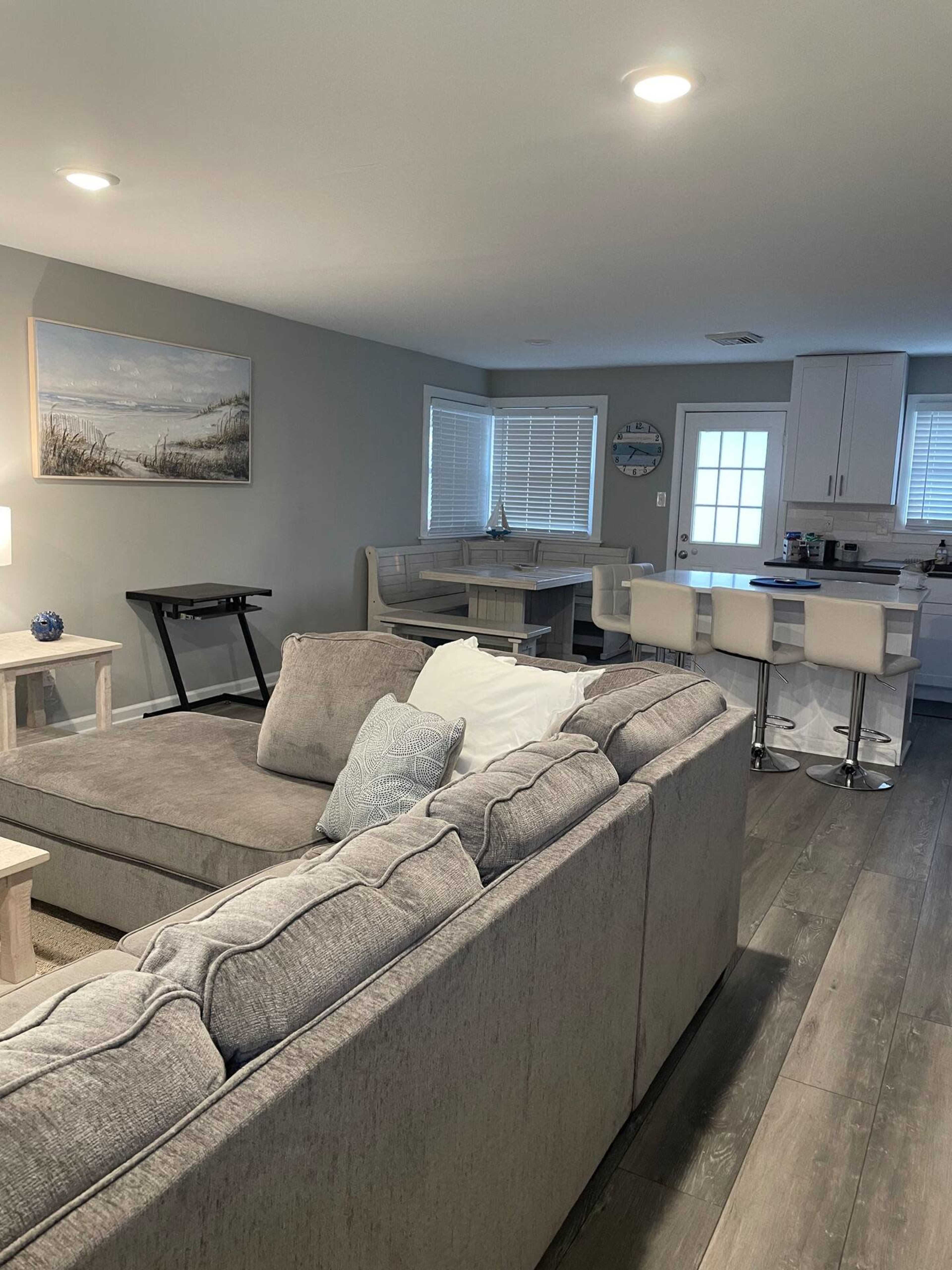 A gray sectional sofa faces a dining area with a table and chairs, while a kitchen is visible in the background with white cabinetry.