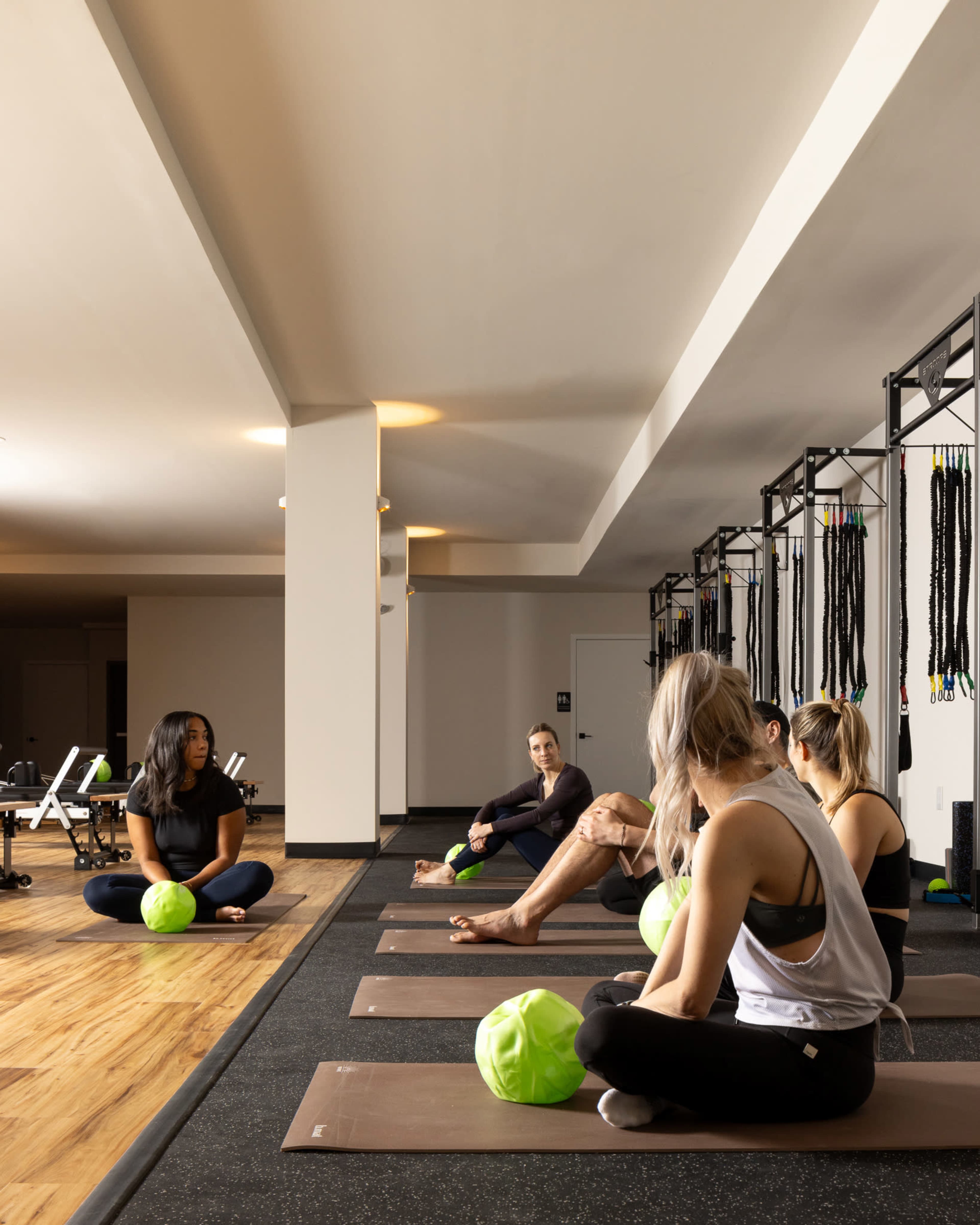 A group of six individuals is seated on yoga mats in a fitness studio, with green exercise balls positioned in front of them and workout equipment visible in the background.
