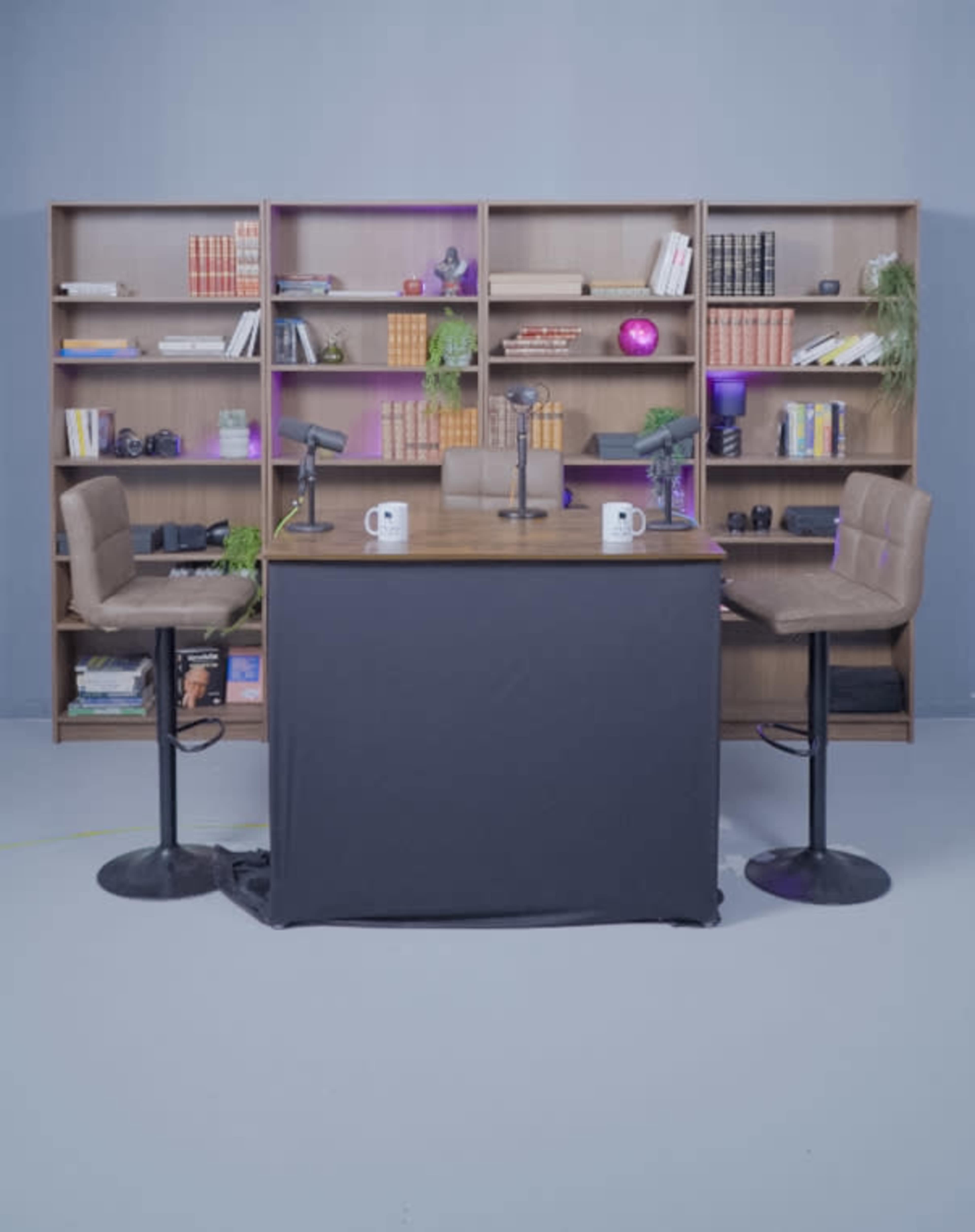 A podcasting or interview setup features a table with two microphones and four bar stools, situated in front of a wall of bookshelves filled with various items.