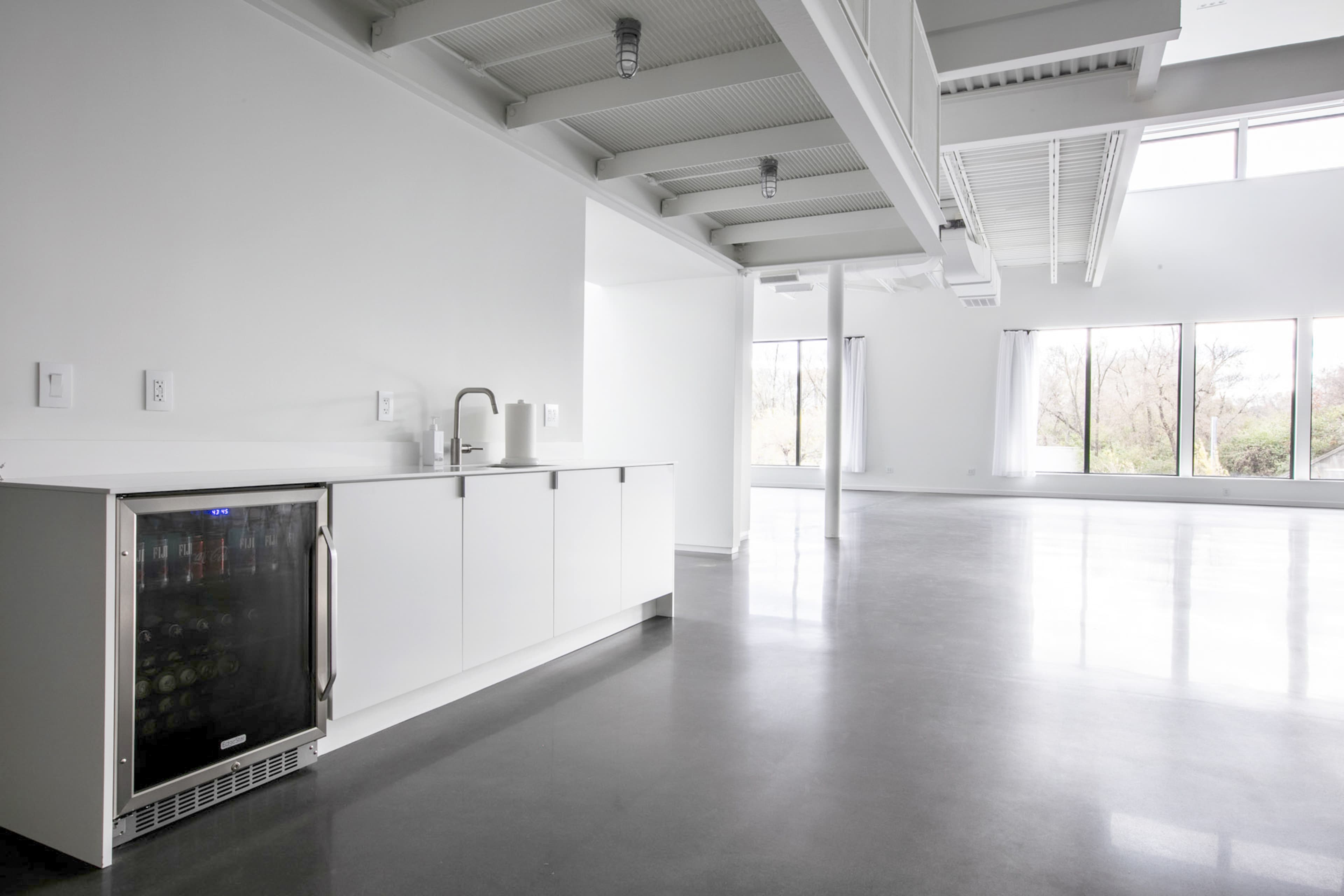 A minimalist, open-concept interior with a small kitchen area featuring a black refrigerator and a countertop, illuminated by large windows that provide natural light.
