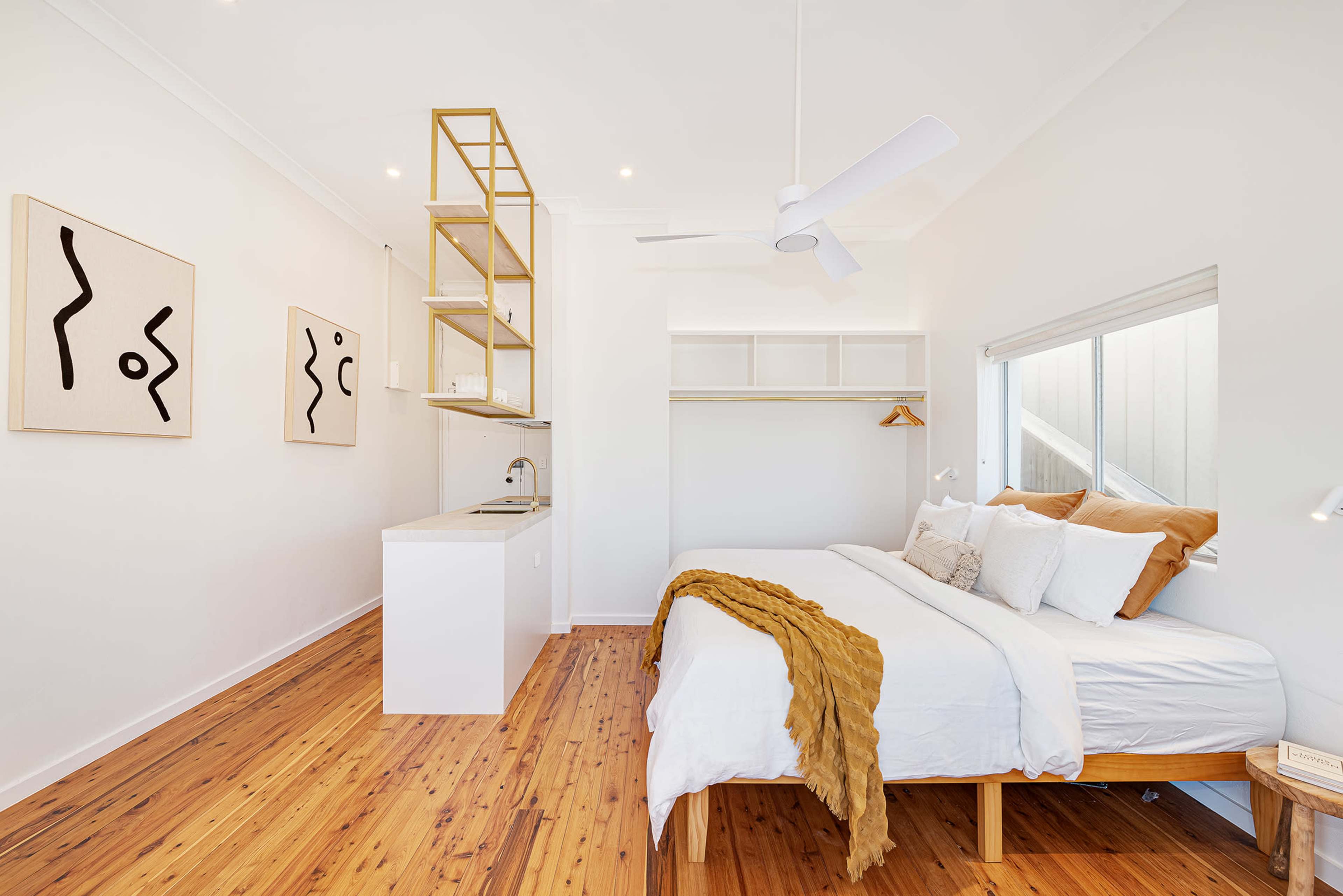 A bright, modern bedroom featuring a large bed with white linens, wooden flooring, abstract wall art, and a small kitchenette against one wall.