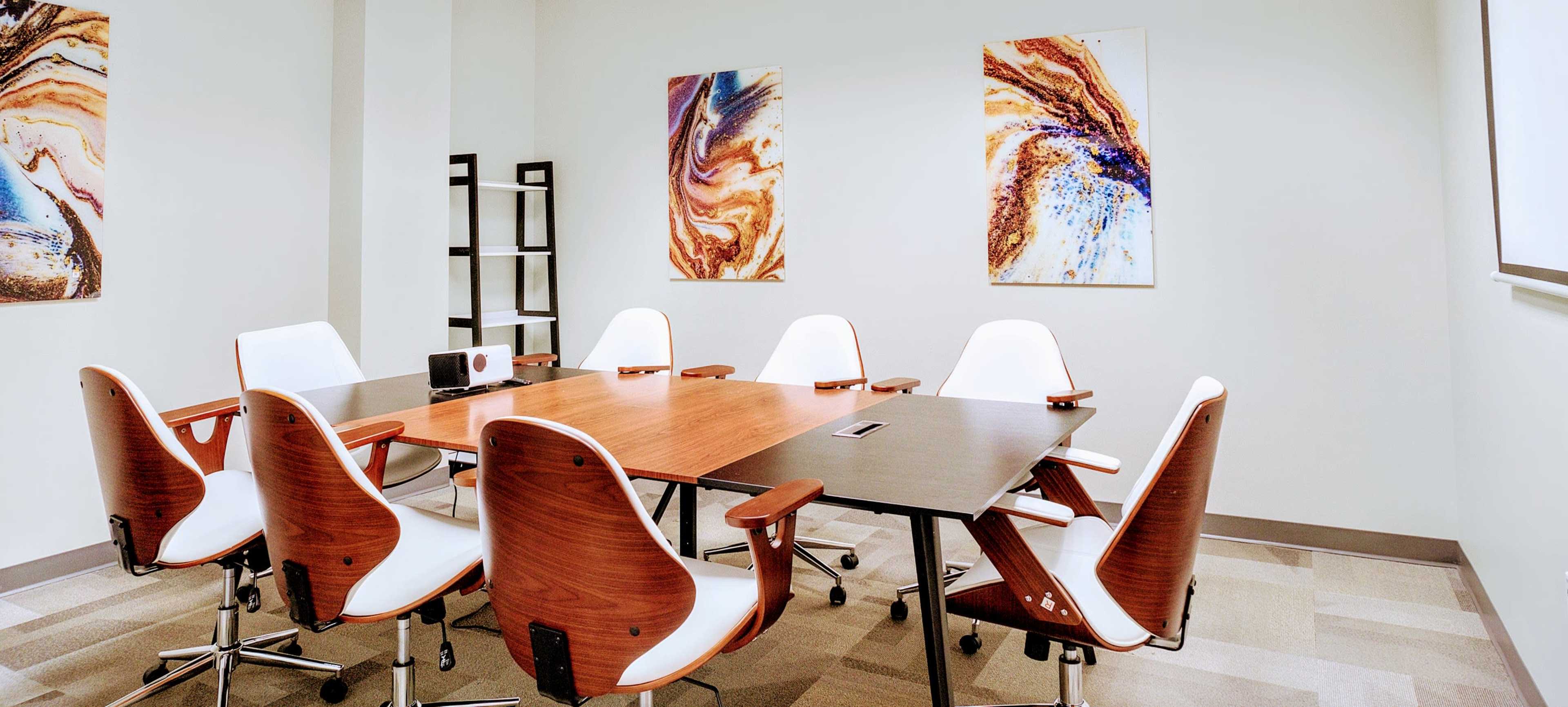 A conference room features a wooden table surrounded by white chairs, with abstract art on the walls.
