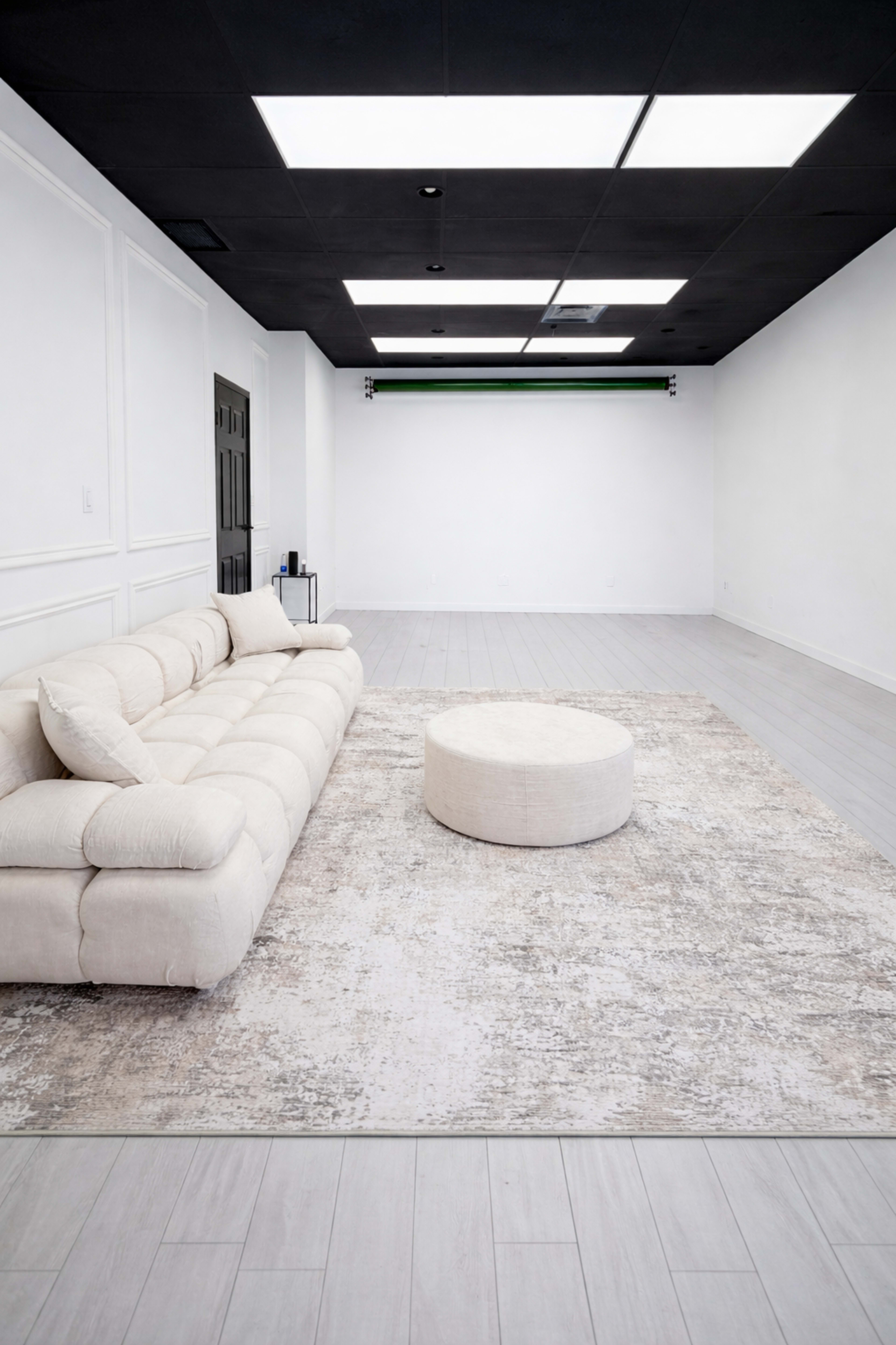 The image shows a minimalist room featuring a light-colored sectional sofa, a circular ottoman, and a large area rug on a wooden floor.