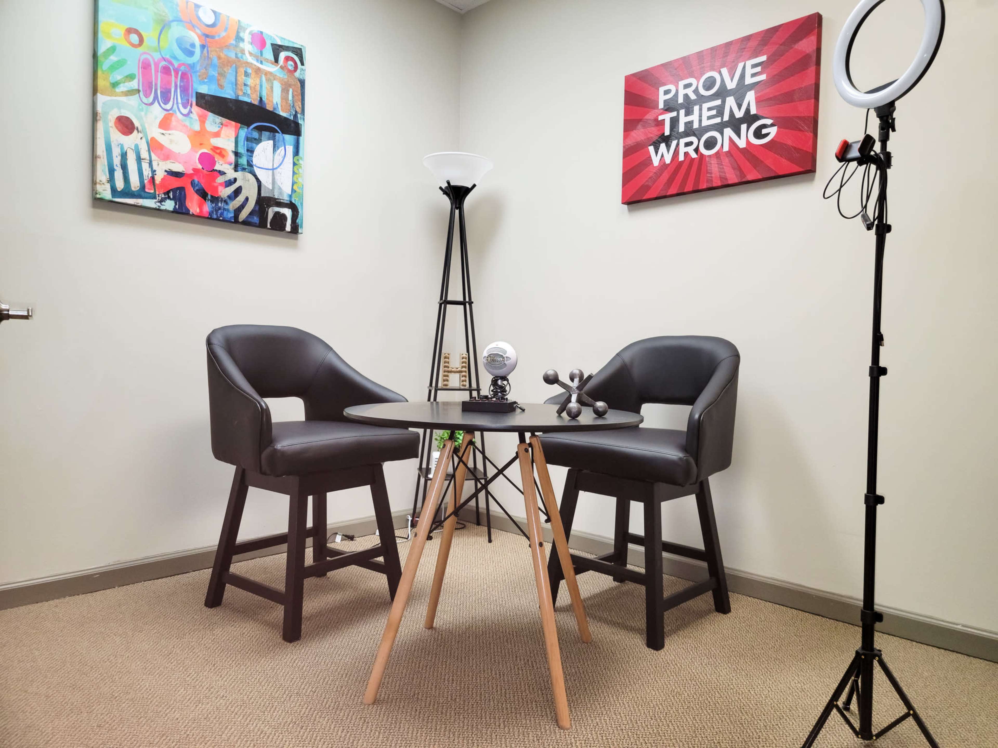 A small office space with two black chairs facing a round table, decorated with art on the walls and a ring light nearby.
