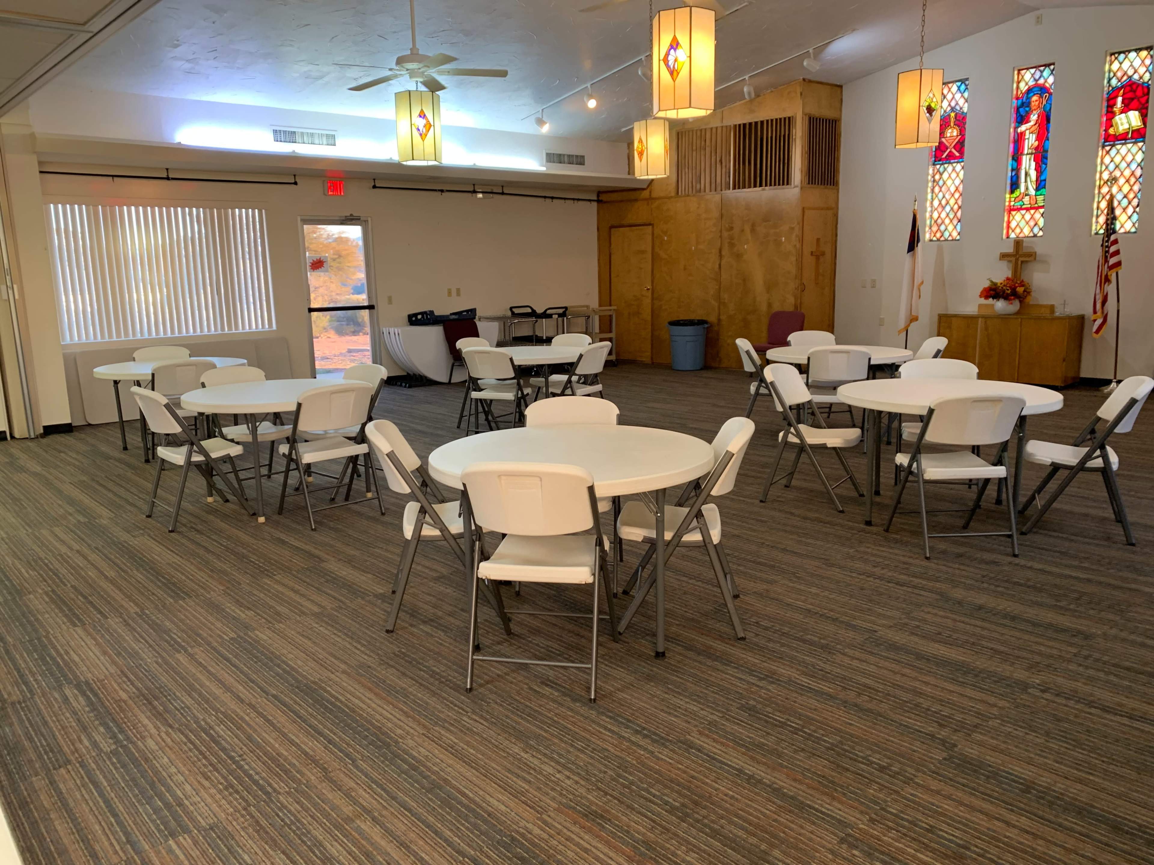 The image shows a spacious room with several round tables and white folding chairs, featuring stained glass windows and a wooden stage area.
