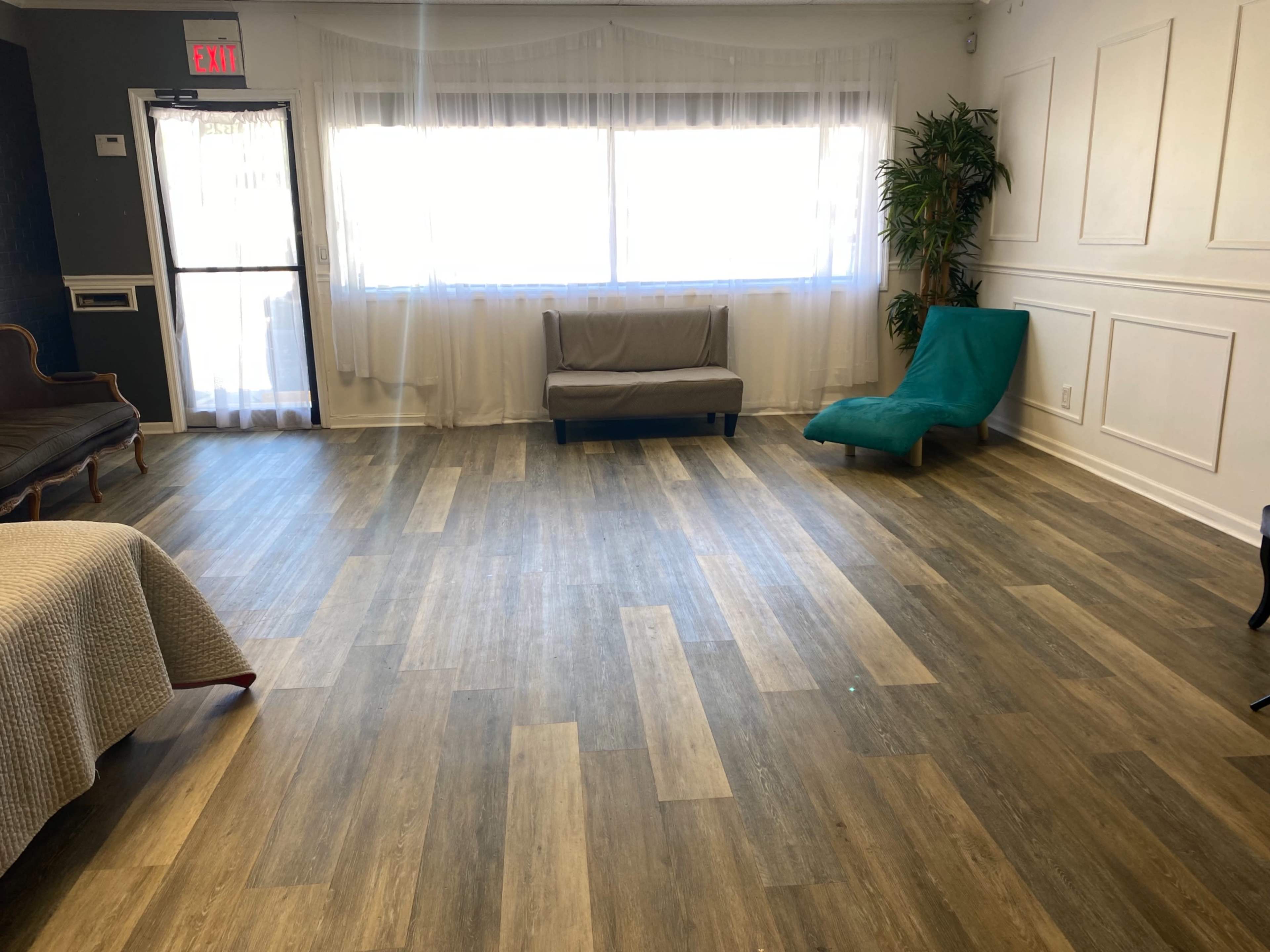 A spacious room with hardwood-style flooring, featuring a window with sheer curtains, a couch, and a green lounge chair.