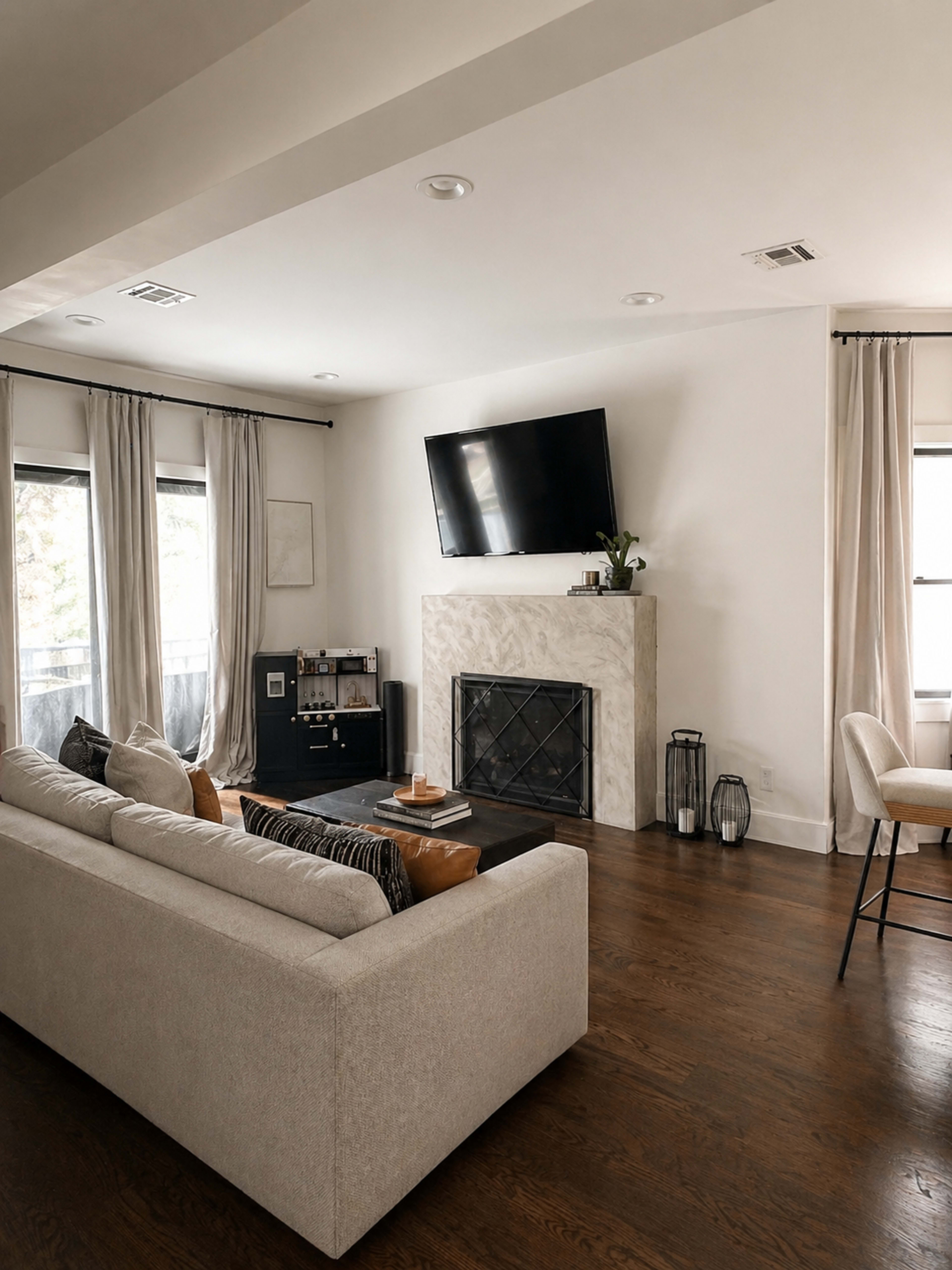 The image shows a modern living room with a large beige sofa facing a wall-mounted television above a marble fireplace, surrounded by large windows with light curtains.