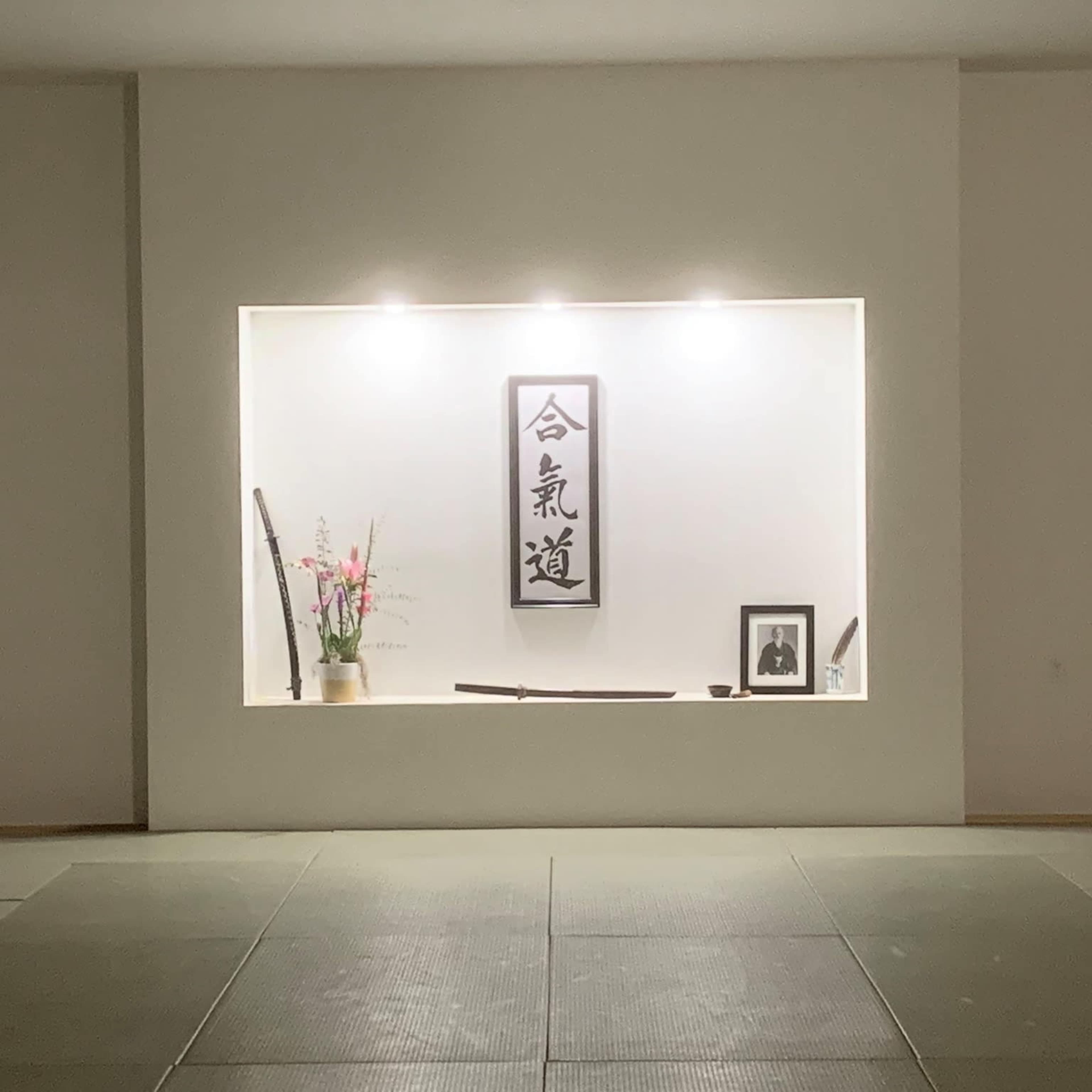 A minimalist display features a wall niche with a calligraphic scroll, a potted plant, a framed photograph, and two traditional Japanese swords.