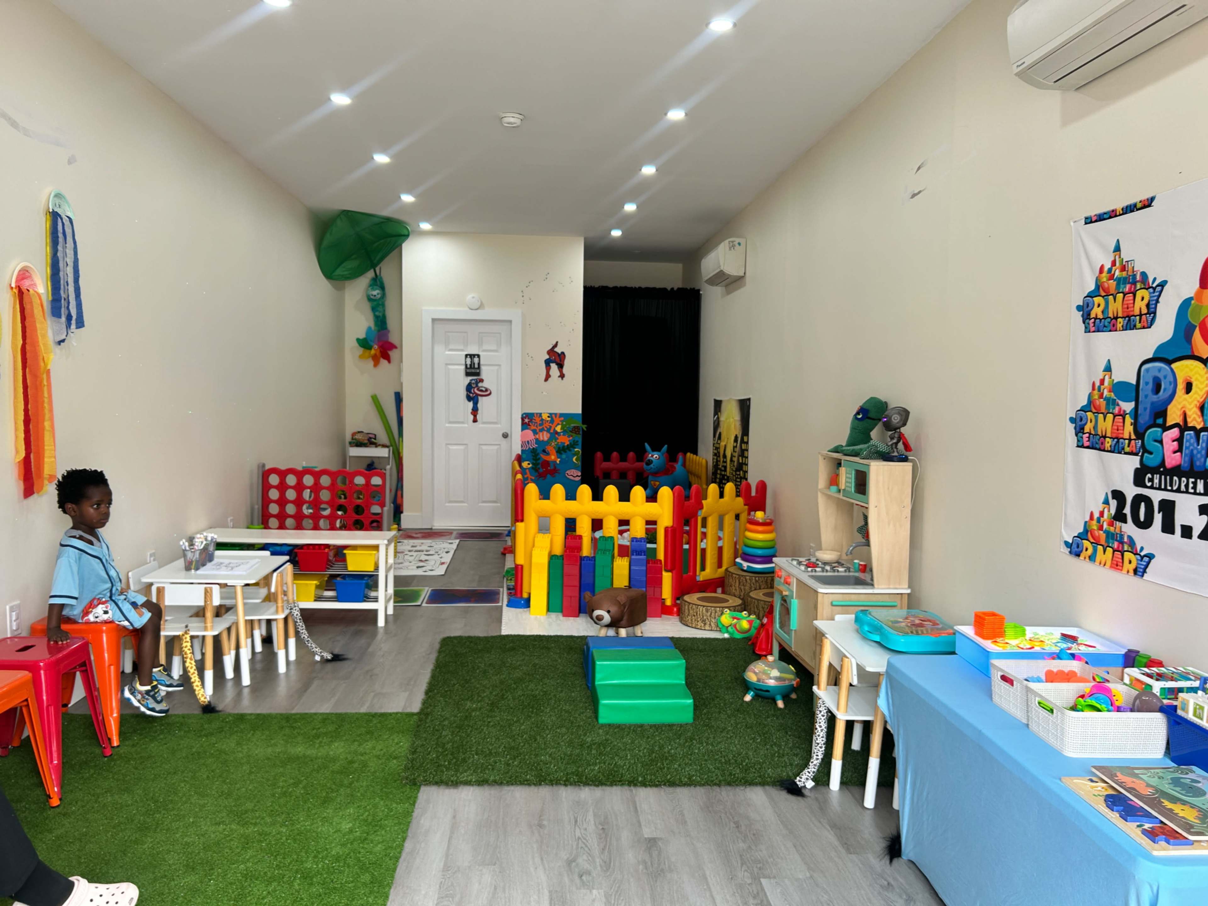The image shows a colorful playroom with children's tables and chairs, a fenced play area, and various toys spread across the floor.