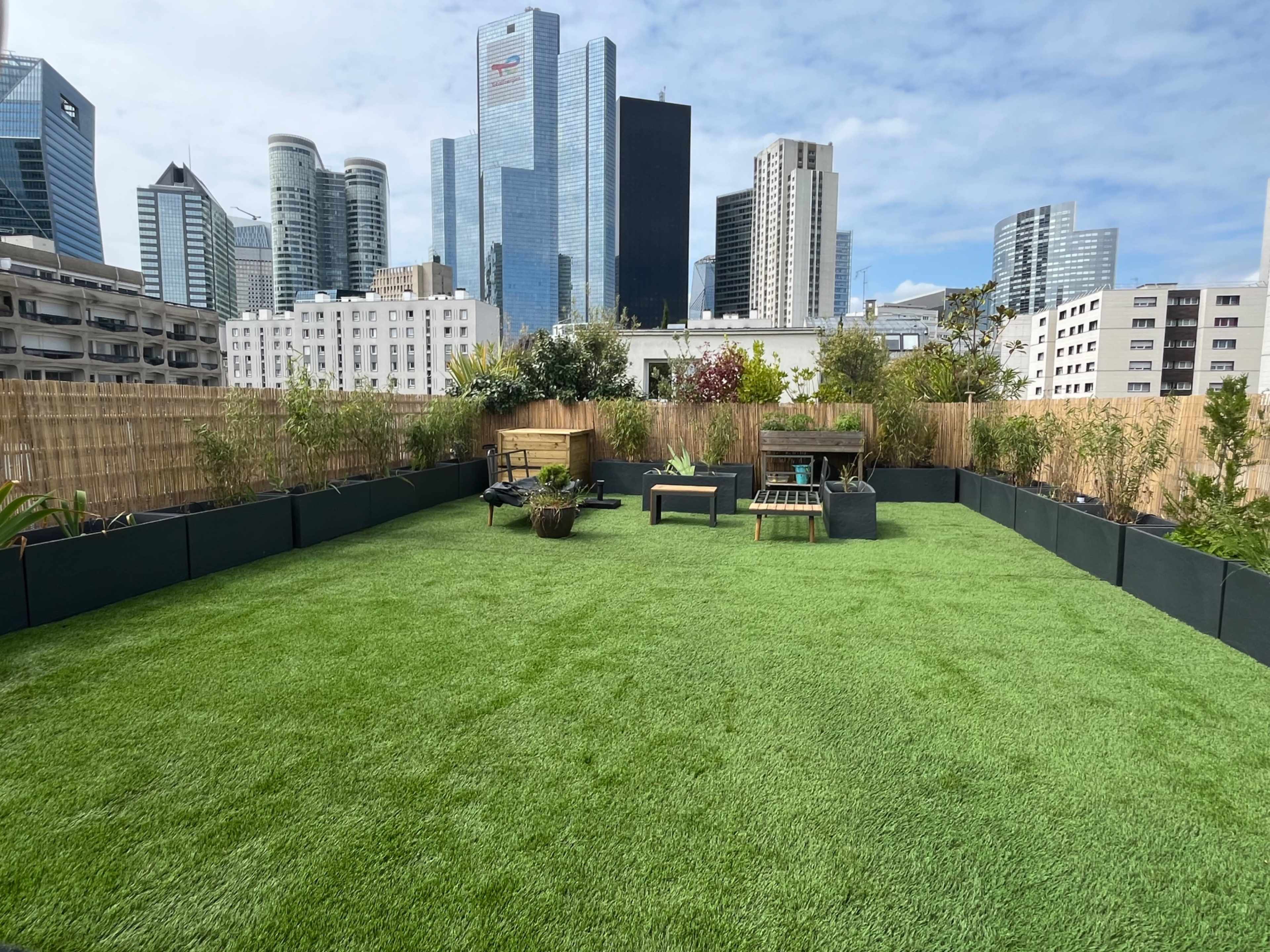 The image shows a rooftop garden with artificial grass, featuring seating areas and surrounding greenery, set against a city skyline.