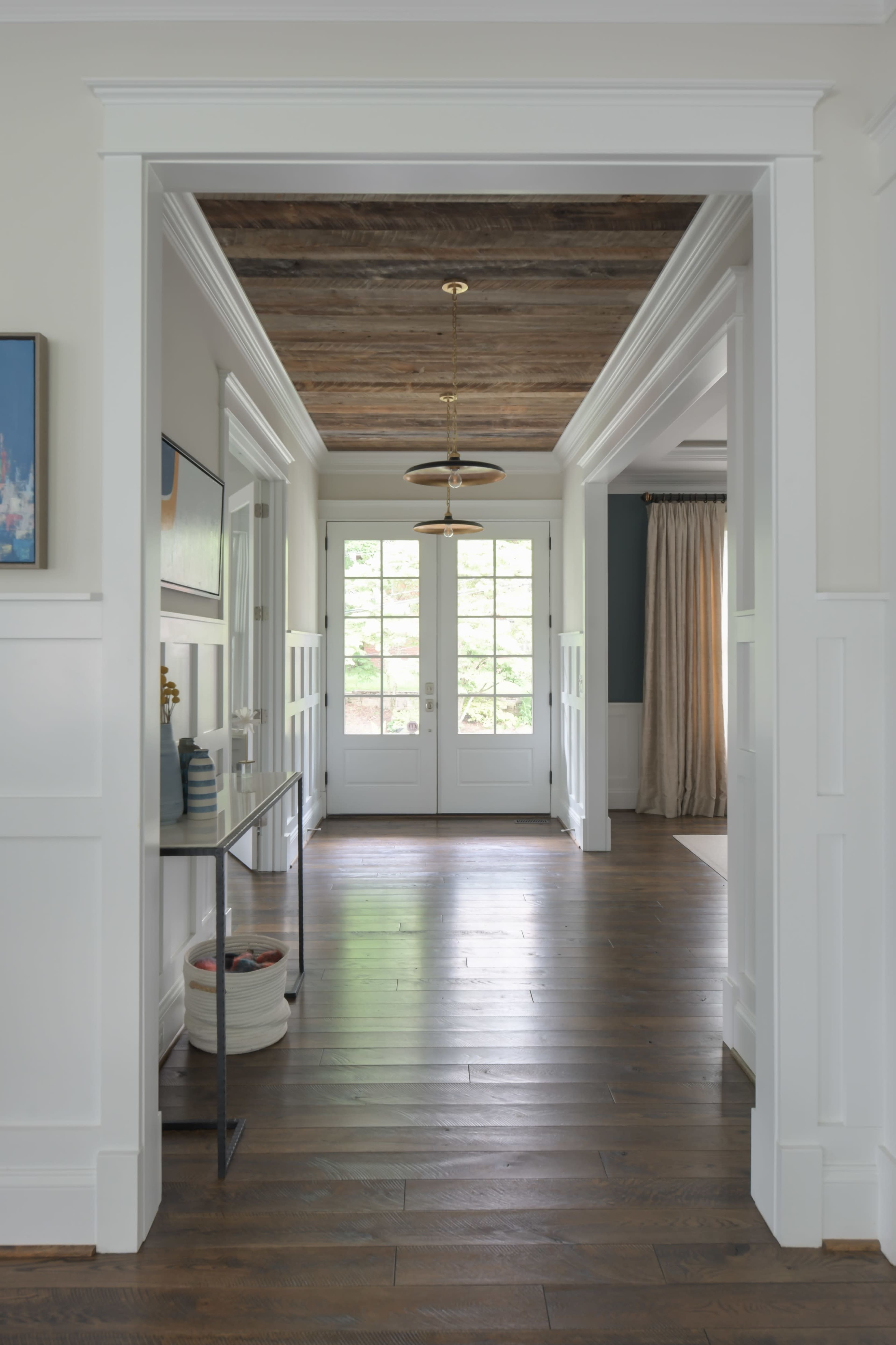A well-lit hallway features a console table and double doors leading to a patio, bordered by paneled walls and a wood ceiling.