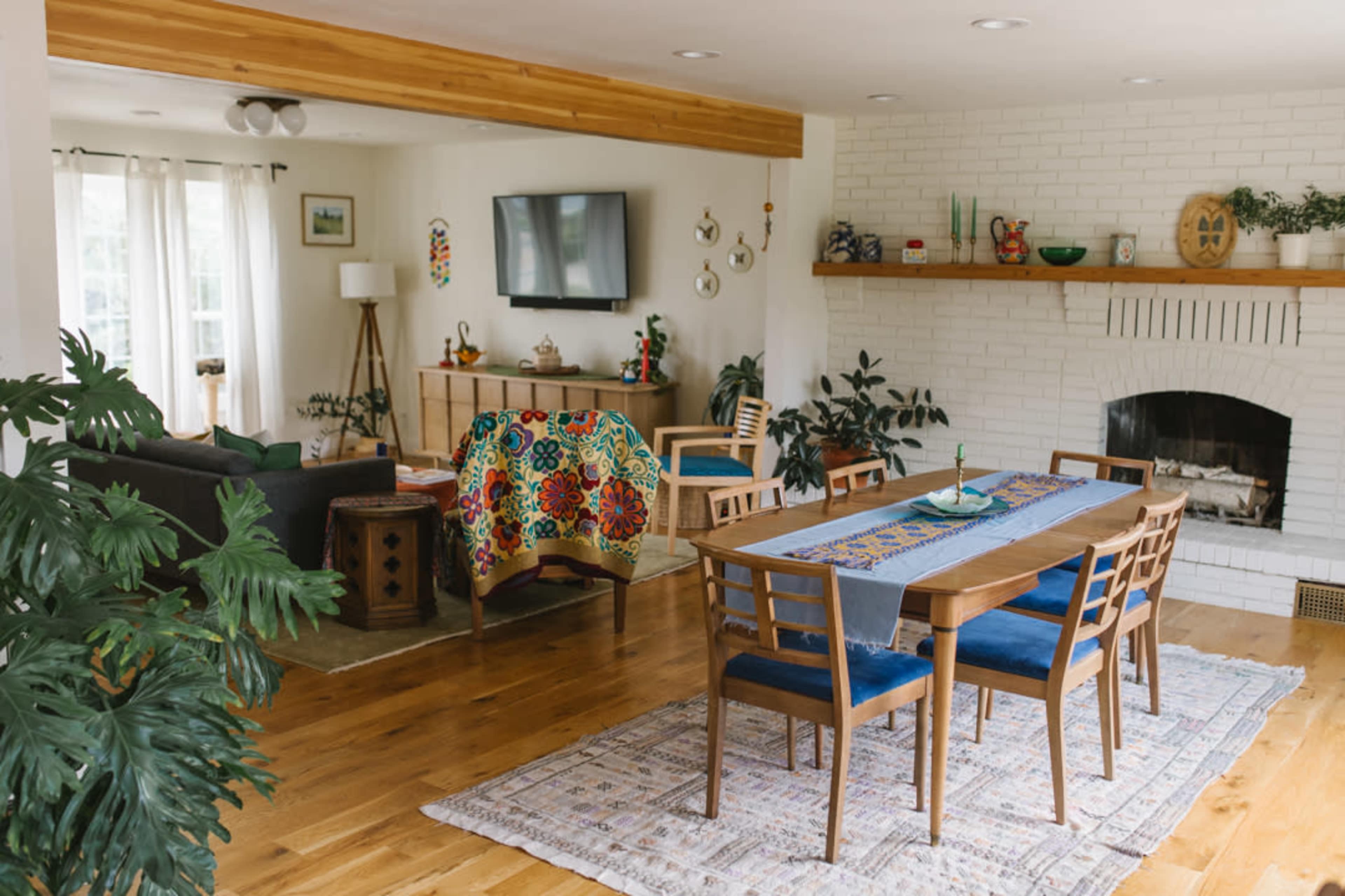 The image shows a modern living and dining area featuring a wooden dining table with chairs, a patterned table runner, a comfortable sofa, and a fireplace with decorative plants and artwork on display.
