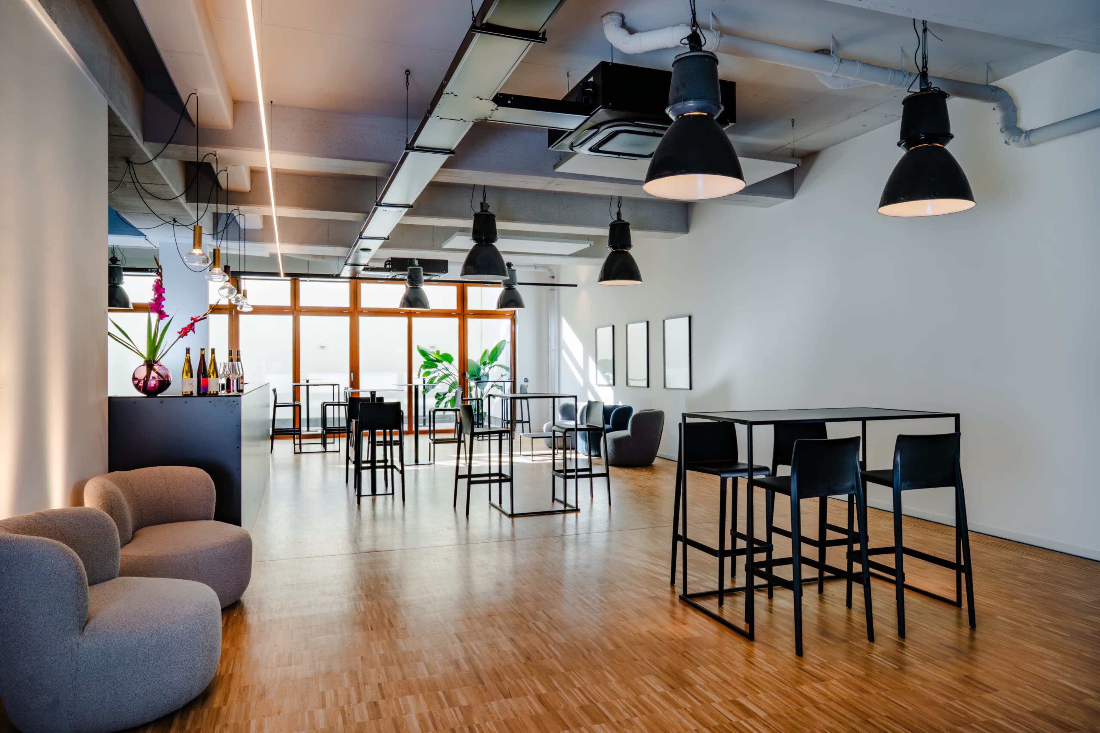 The image shows a modern, open space with wooden flooring, featuring several tall tables and black chairs, along with a gray seating area and large windows allowing natural light in.