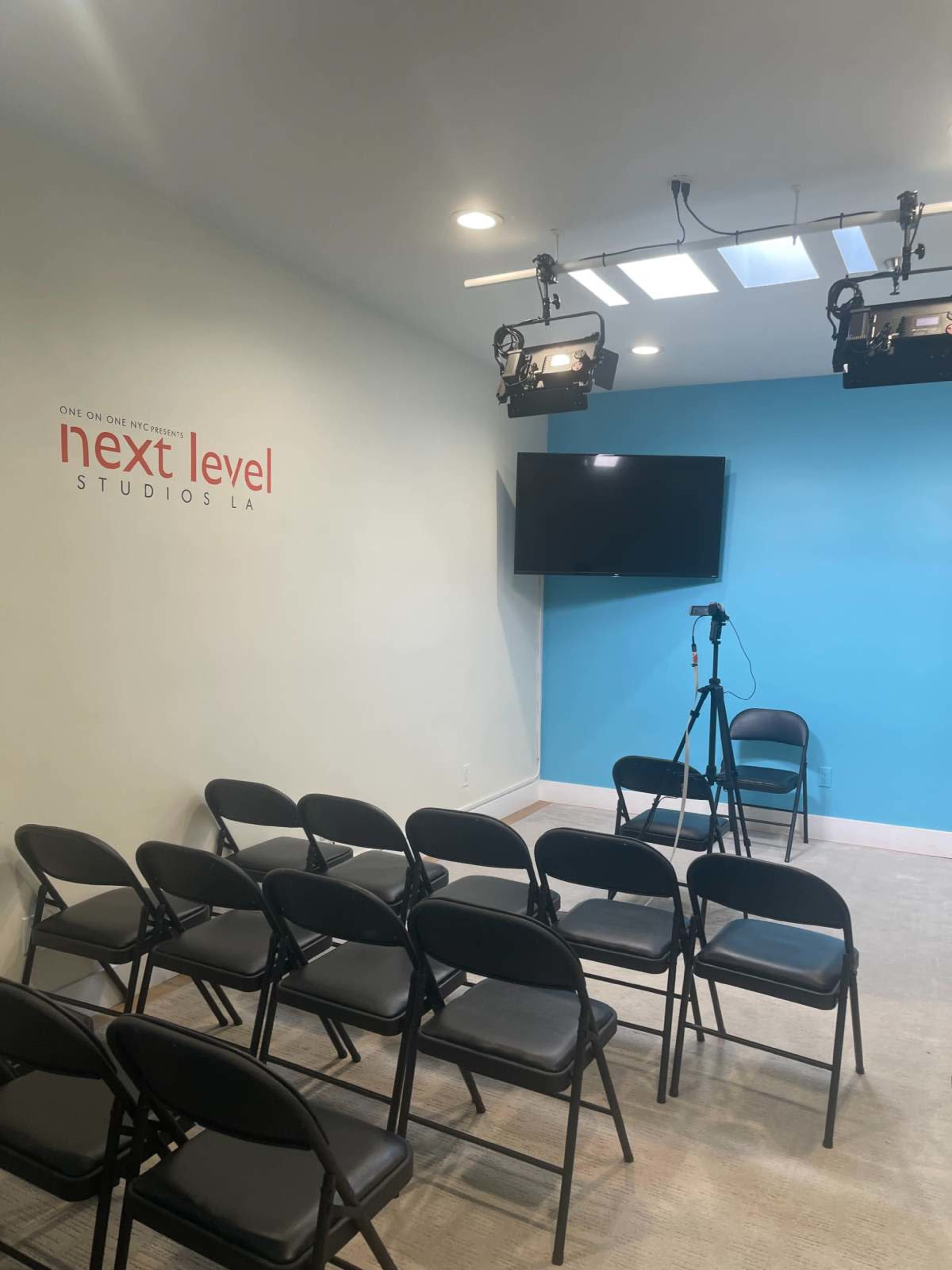 The image shows a small room with blue walls, several black folding chairs arranged in rows, a mounted television screen, and studio lighting overhead.