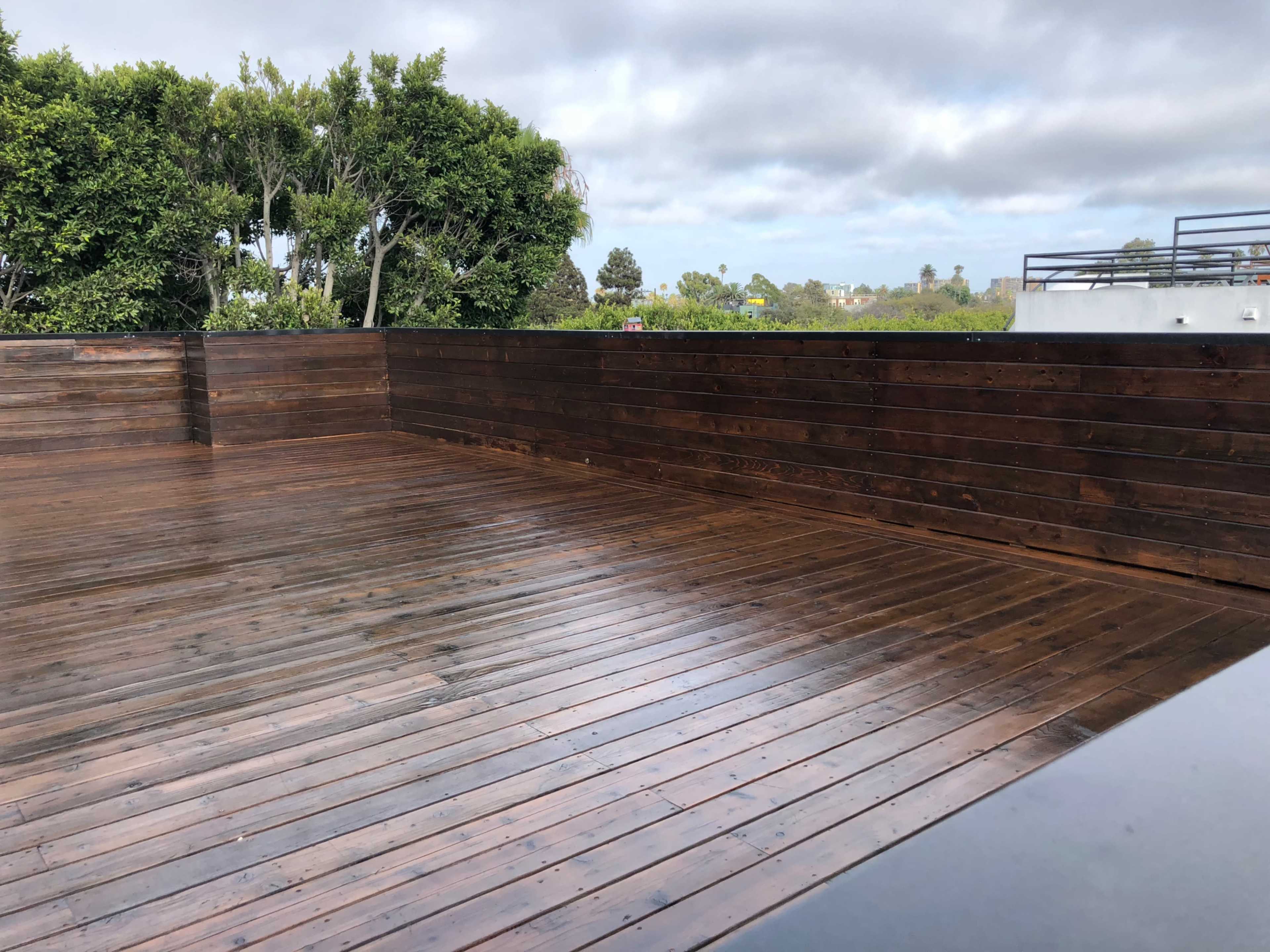 The image shows a wooden deck with a dark finish, surrounded by green foliage under a cloudy sky.