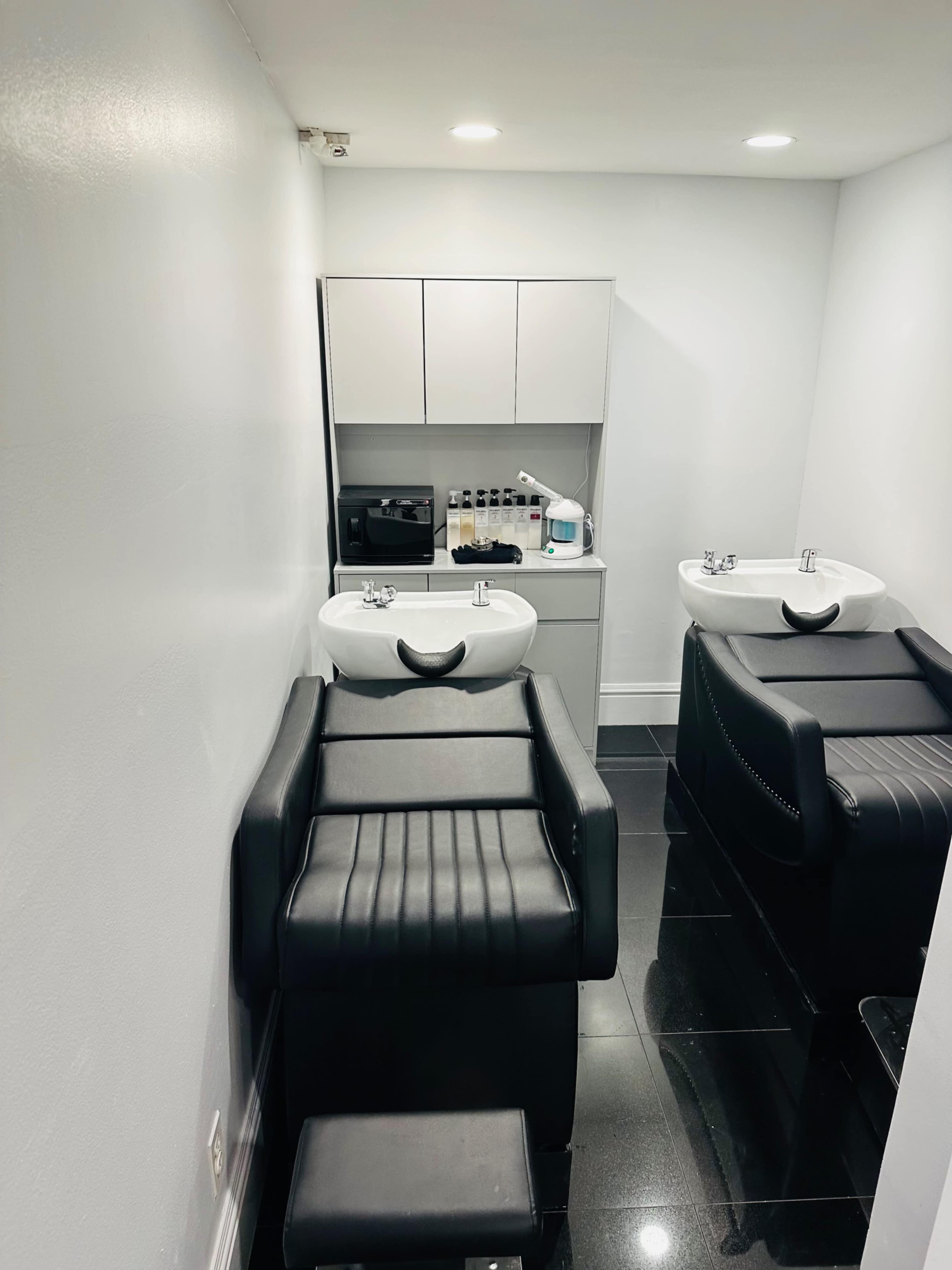 The image shows a minimalistic salon space featuring two black treatment chairs and a wall-mounted cabinet with various products.