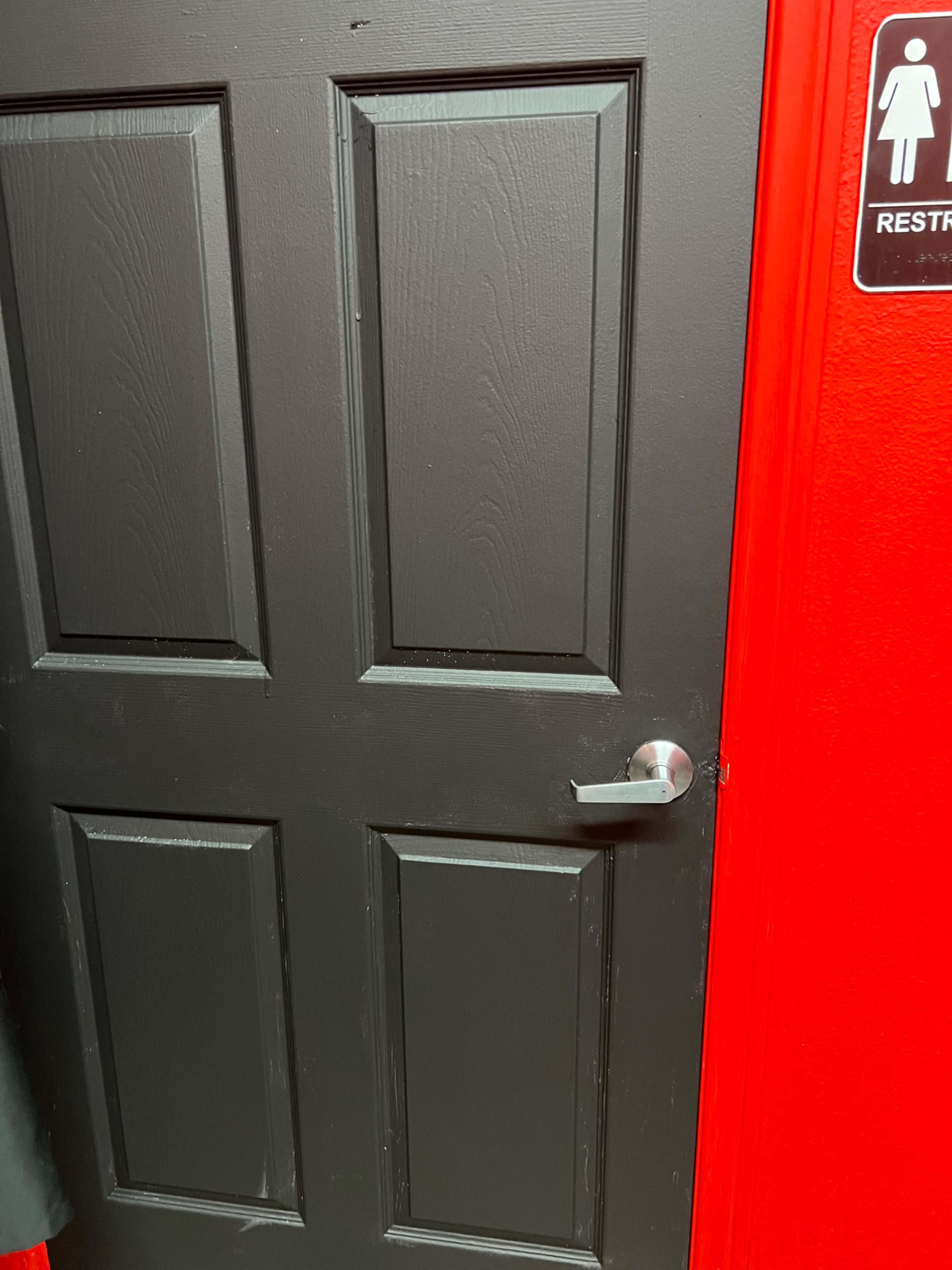 A black paneled door with a silver handle is positioned next to a bright red wall featuring a restroom sign.