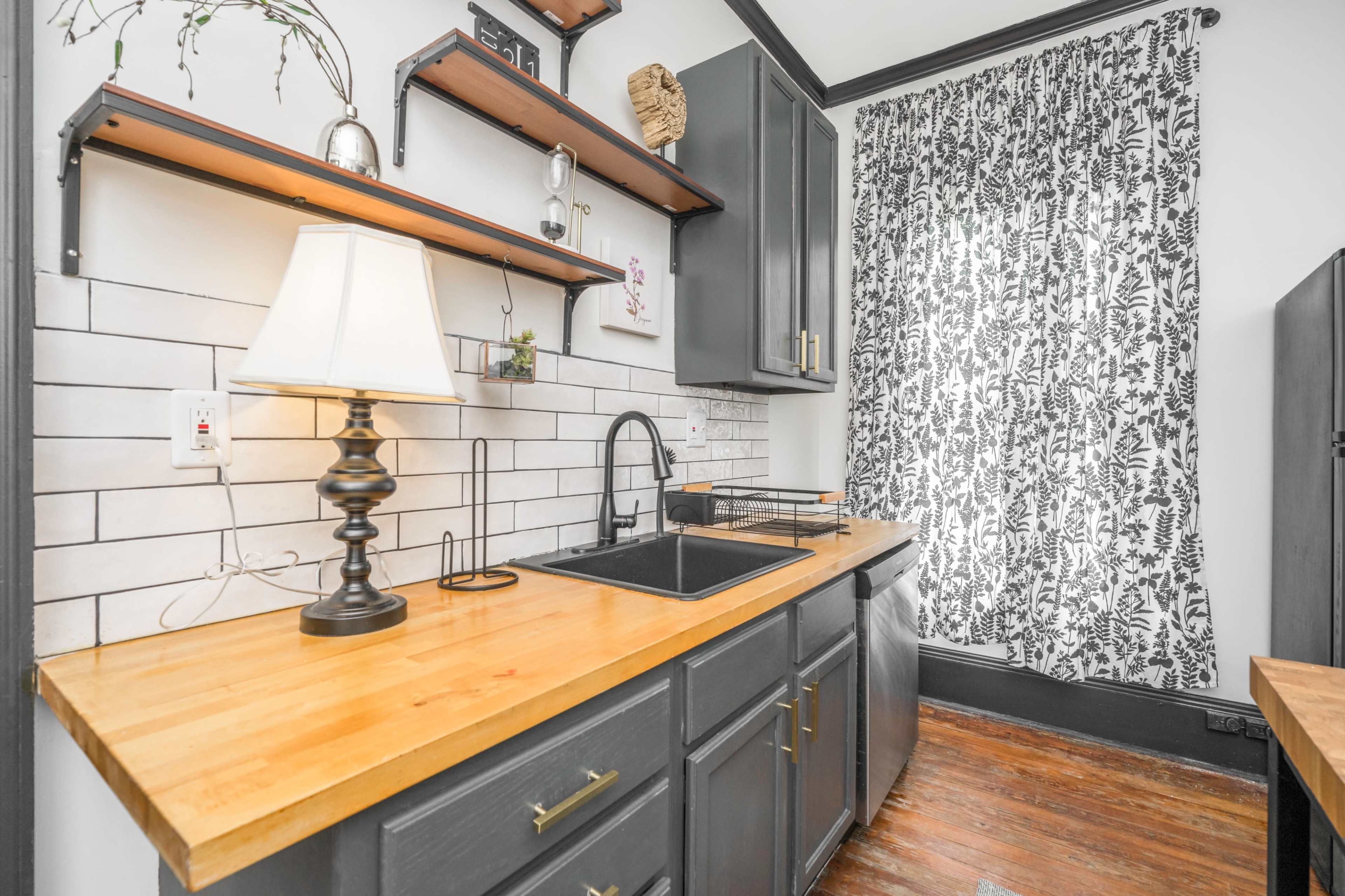 The image shows a modern kitchen with dark cabinetry, open shelving, a wooden countertop, a black sink, and a large patterned curtain.