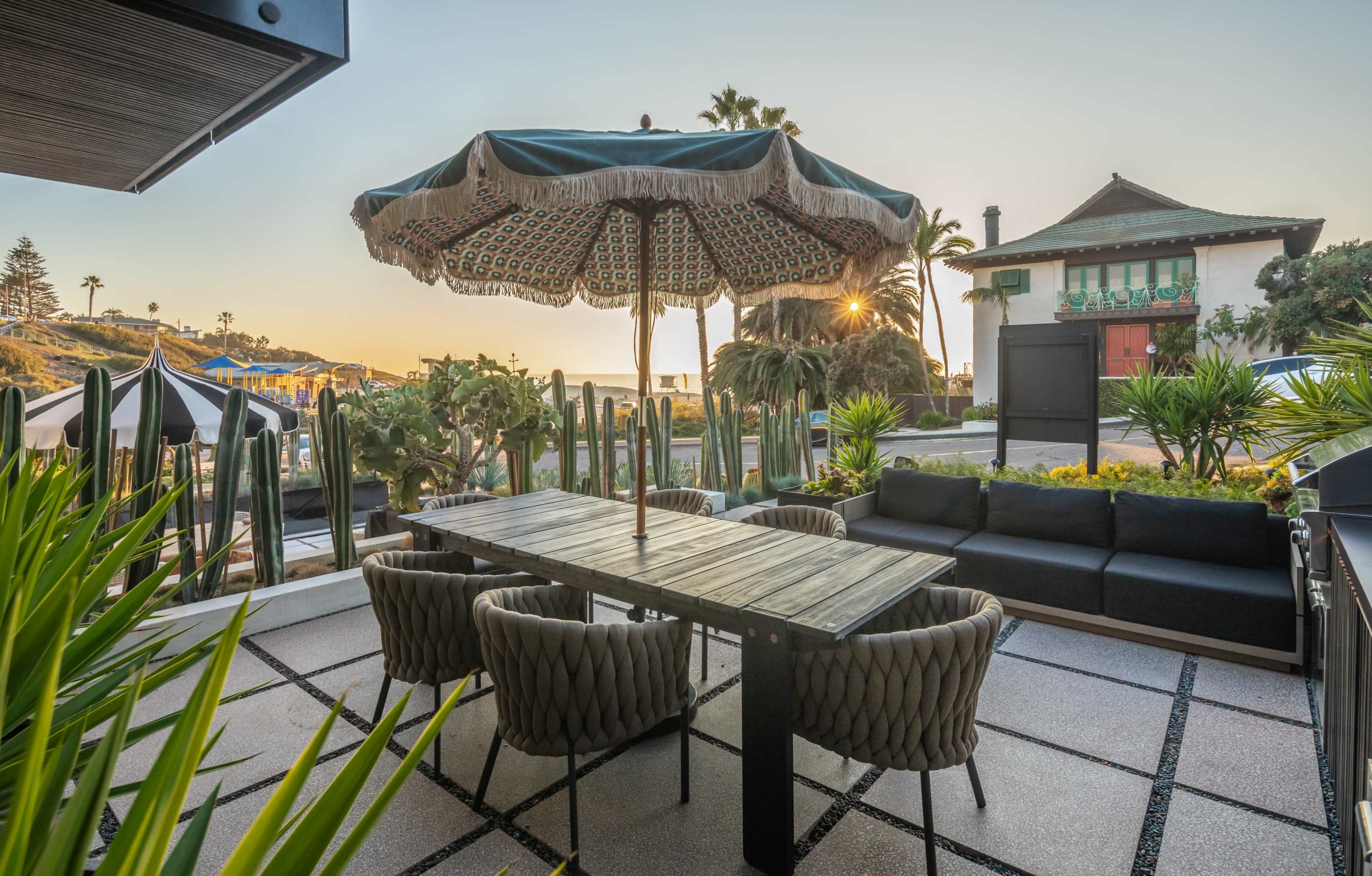 A patio area features a large umbrella over a dining table surrounded by woven chairs, with palm trees and a building in the background illuminated by sunset.